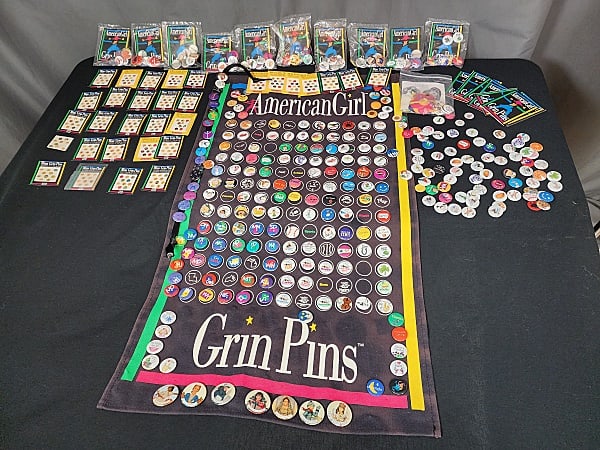 Large Group of American Girl Doll Grin Pins and More: American Girl Doll Grin Pins Minis, and banner. Group includes a banner with 53, 1'' grin pins and 7, 1.5'' pins. There is a black ribbon with 18 grin pins with an additional loose 72 grin pins, and 1