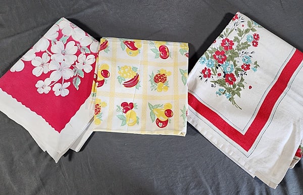 3 Vintage Printed Tablecloths-Fruit and Flowers: Group includes white cotton with white and grey dogwood flowers against a red frame, measures 44"x47", has spots, fading & storage discoloration. White cotton with red and yellow fruit, measures 39"x4