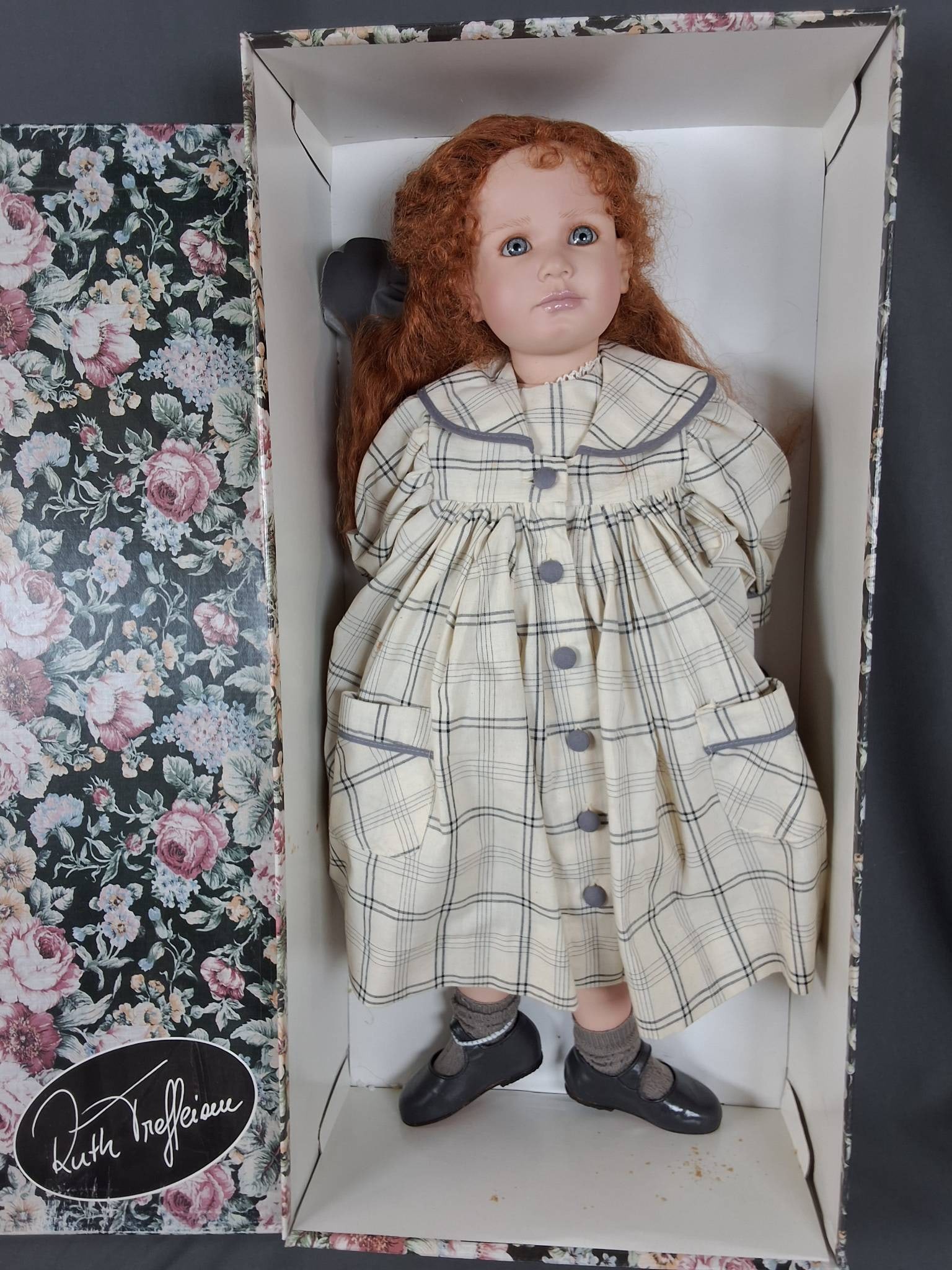 Lisa by Ruth Treffeisen: Lisa by Ruth Treffeisen stands 23 inches tall and features a PVC head with PVC limbs. She wears a plaid cotton dress with soft tones in its pattern and has white bloomers underneath, complemented by l