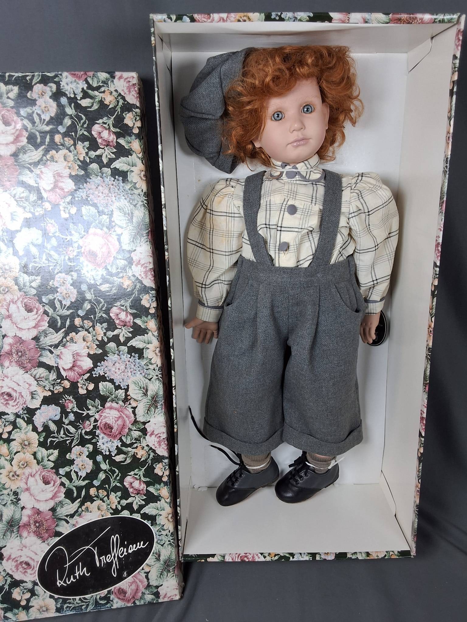 Alex by Ruth Treffeisen: Alex by Ruth Treffeisen stands 23.5'' tall and features a PVC head and limbs. He is dressed in a plaid shirt paired with long gray pants held in place by matching suspenders. A gray hat adds to his ou