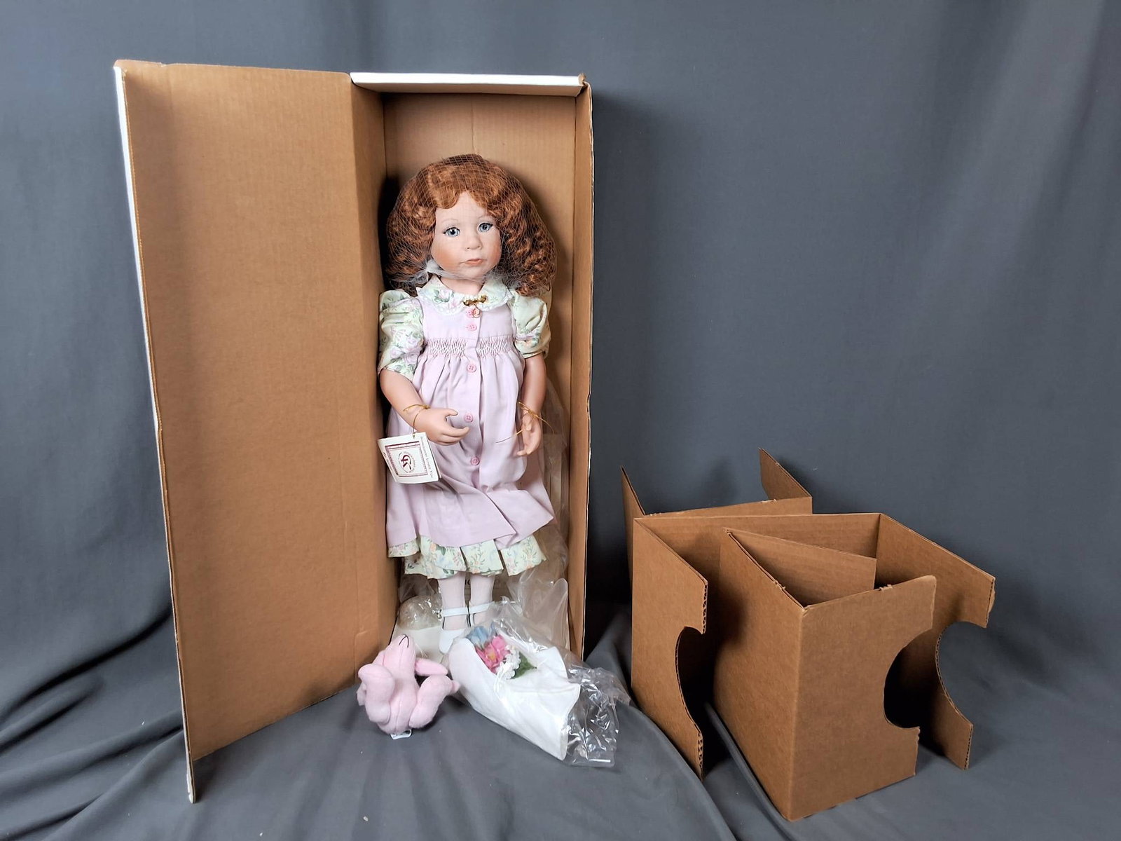 Somebody Loves You by Julie Good-Krueger: Standing at 22 inches tall, this vinyl doll is part of a limited production of 1000. She is dressed in a pastel outfit featuring a light purple pinafore with pink buttons over a floral-patterned dress