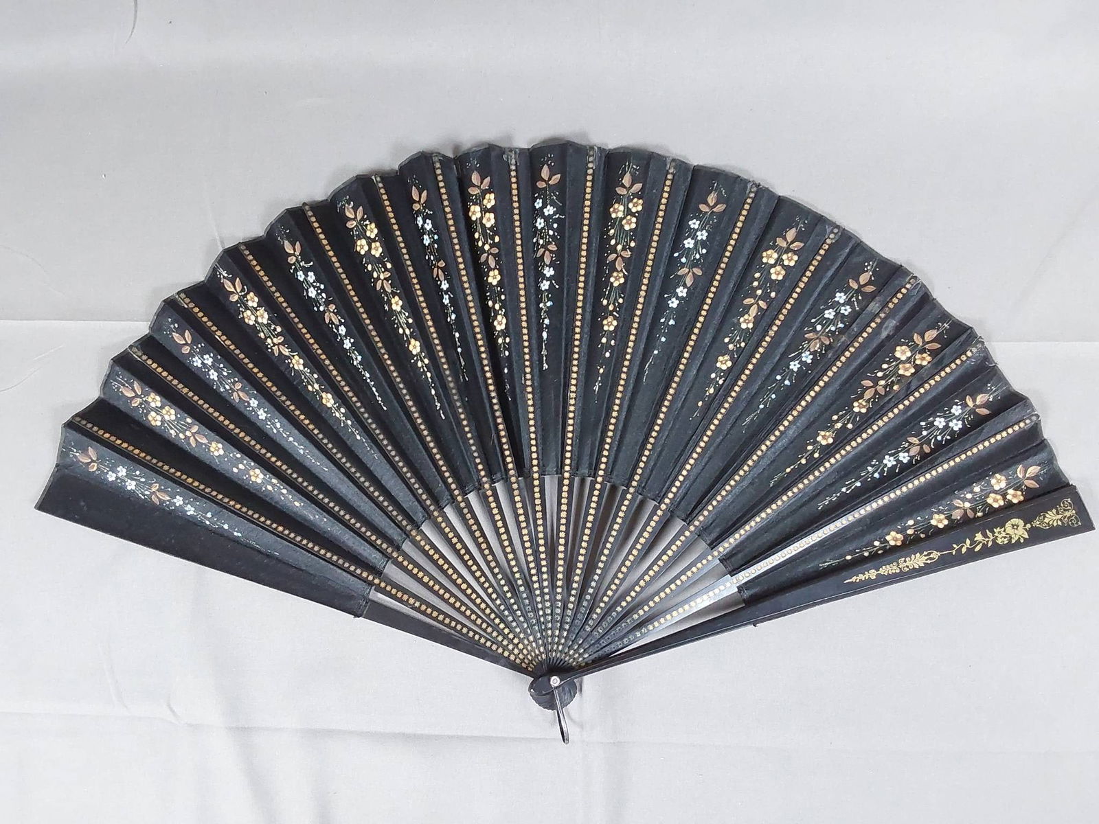 Antique Hand Painted Black Silk Folding Fan: Antique hand painted black silk folding fan featuring hand painted floral sprays on in shades of gold and light blue, complemented by wood guards and ribs with gold tone embossed star and floral motif