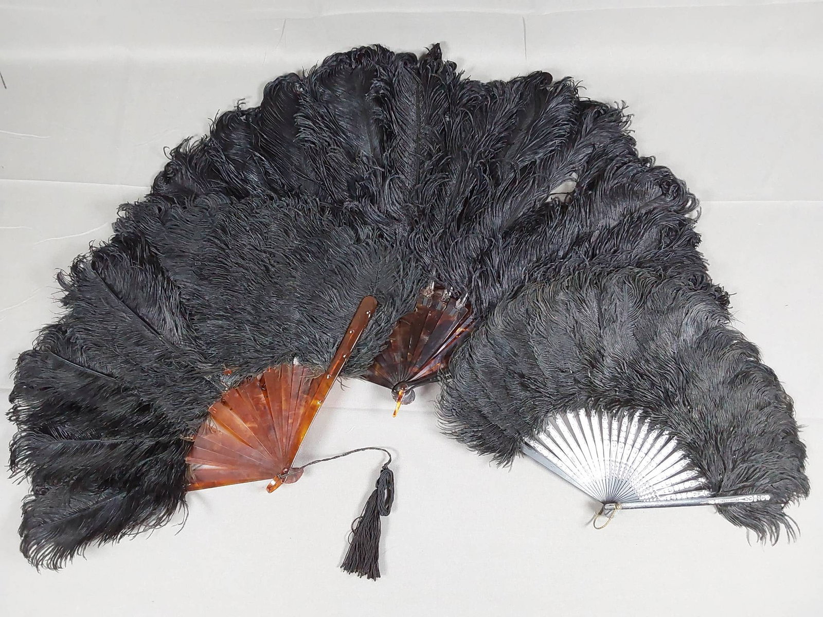 3 Antique Black Ostrich Feather Evening Fans: 3 antique evening fans with black ostrich feathers. The largest fan measures 16 in x 26 in and faux tortoise shell celluloid guards and ribs, with a silver-toned applied ID monogram. The ribbon lacing
