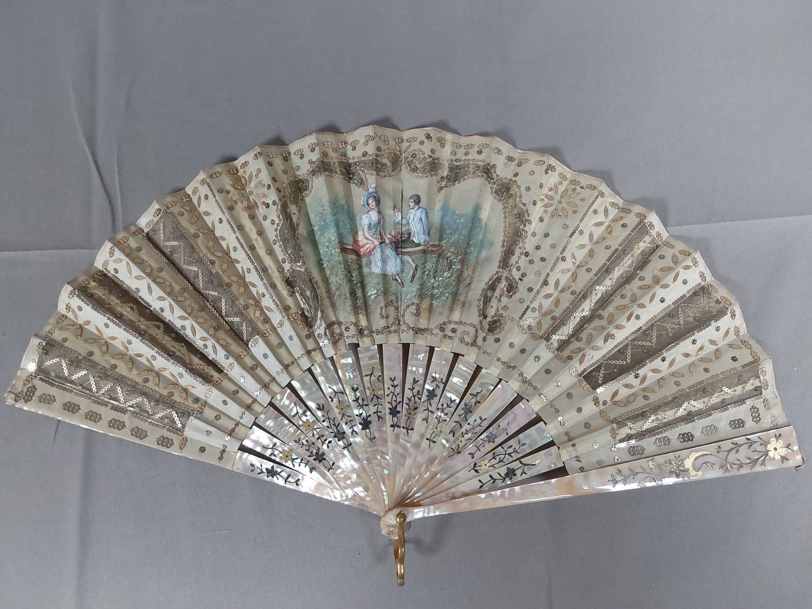 Antique Hand Painted Folding Fan - Mother Of Pearl Guards: Antique hand painted folding fan made of silk, decorated with gold spangles, depicting a romantic scene and decorative transparent areas. Mother of pearl guards and ribs feature intricate floral inlay