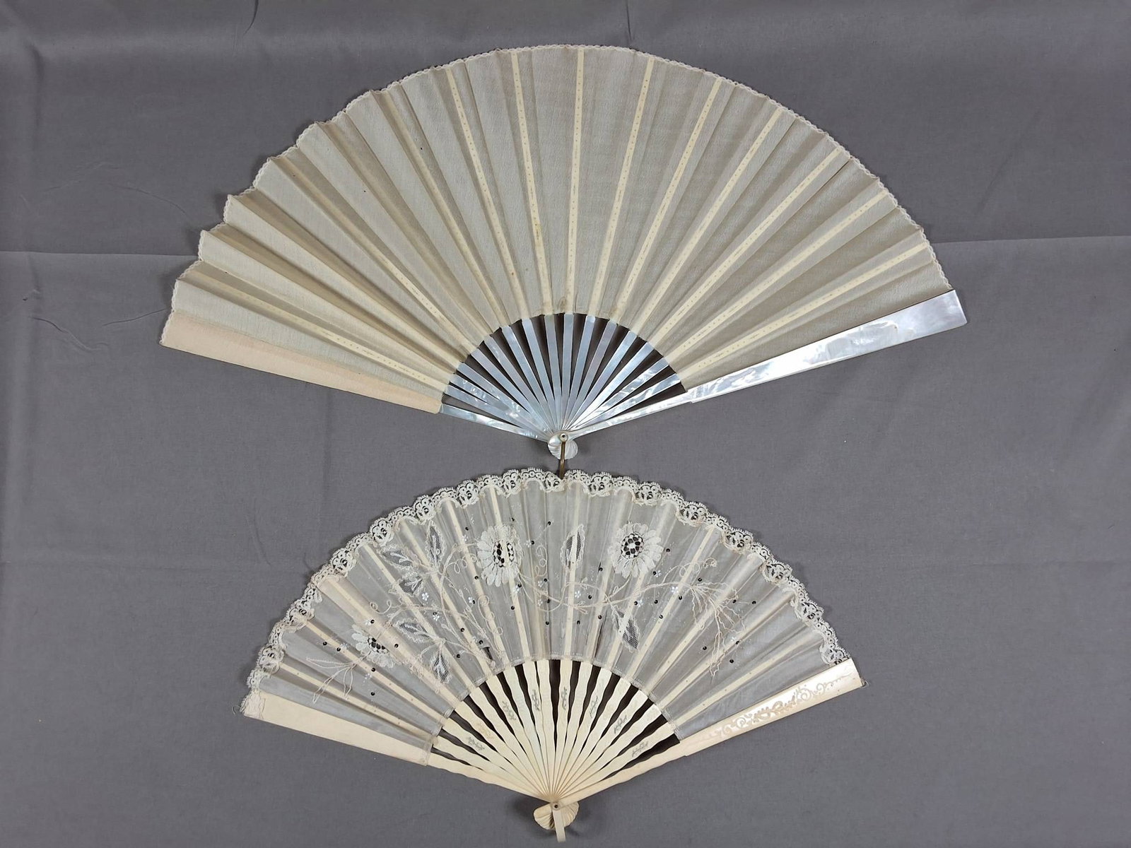 2 Antique Lace Trimmed Silk Folding Fans: Lot includes two silk antique folding fans. The larger fan features a silk leaf with delicate scalloped lace trim, mother of pearl guards and ribs, pierced bone ribs visible on the reverse, a metal lo