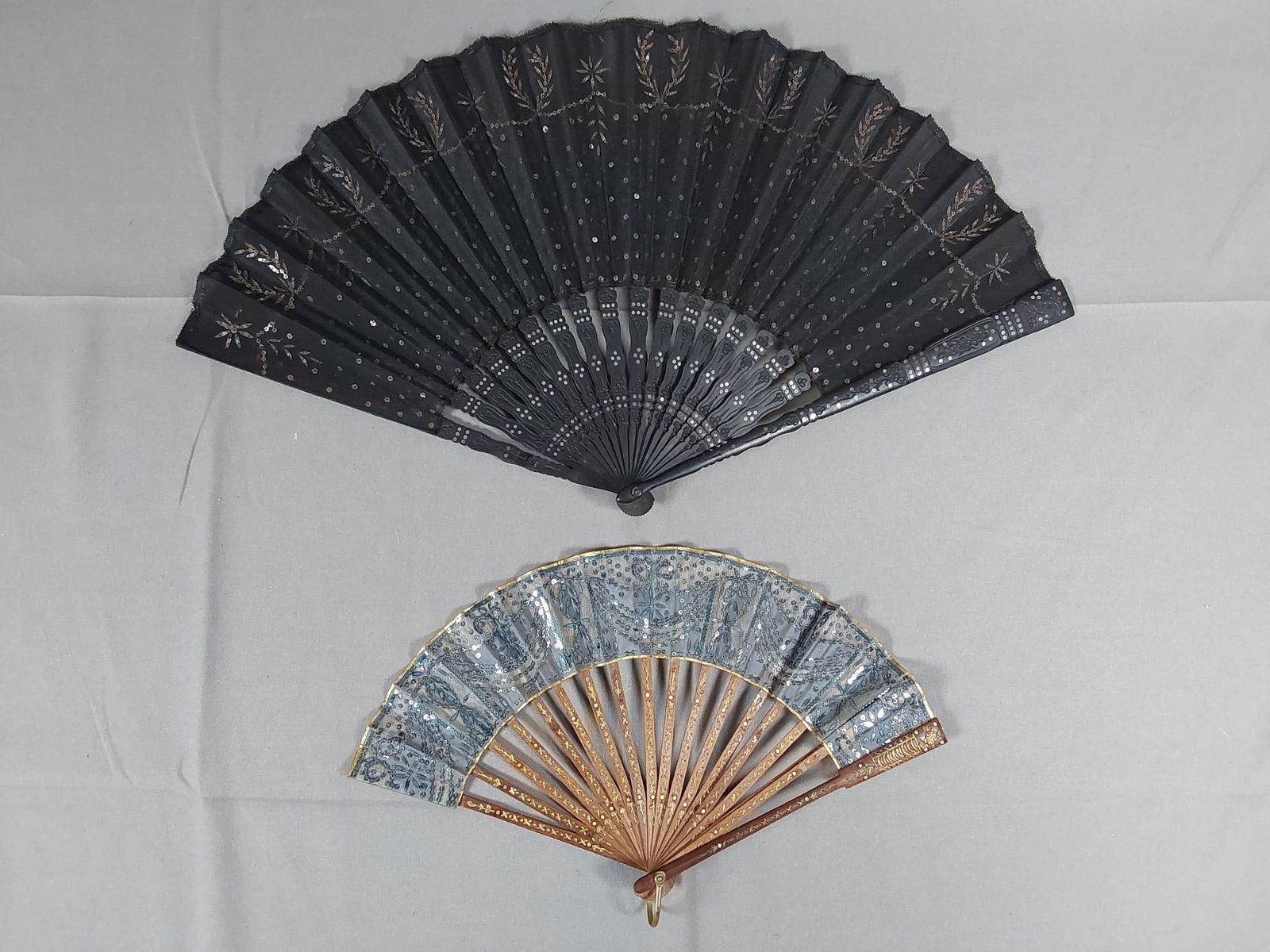 Antique Silk Fans Model Small Blue and Large Black - Metallic Spangles: Two antique silk fans featuring metallic spangles. The smaller fan, measuring 6 1/4" x 11 1/4", is blue with appliqued designs and matching spangles, wood guards and ribs with gold-colored inlay, a go