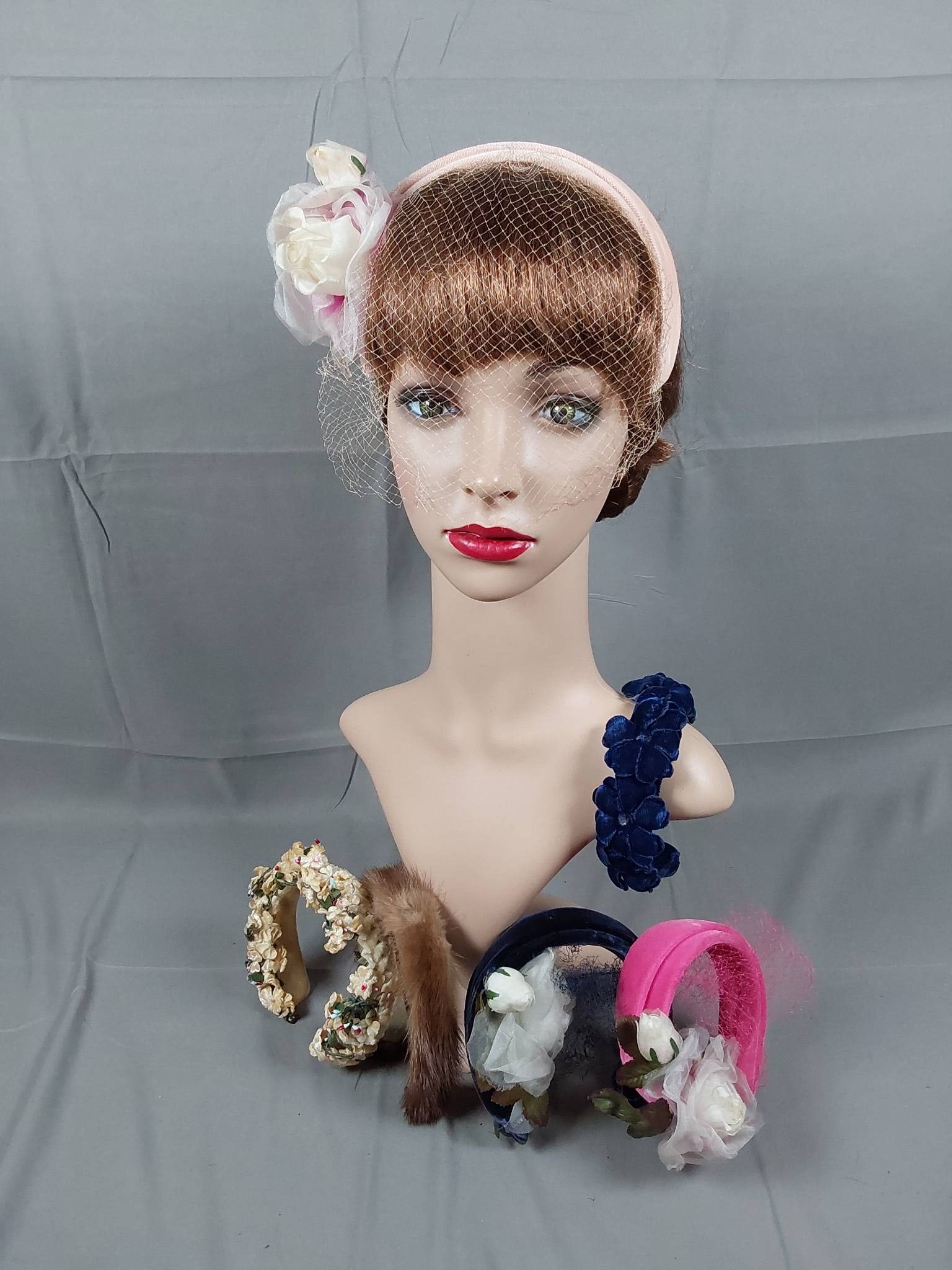 Group of 6 Vintage 1950s Headbands: Group of six vintage 1950s headbands featuring a variety of designs and materials. Includes three velvet-covered headbands in light pink, pink, and navy blue with silk rose embellishments and a matchi