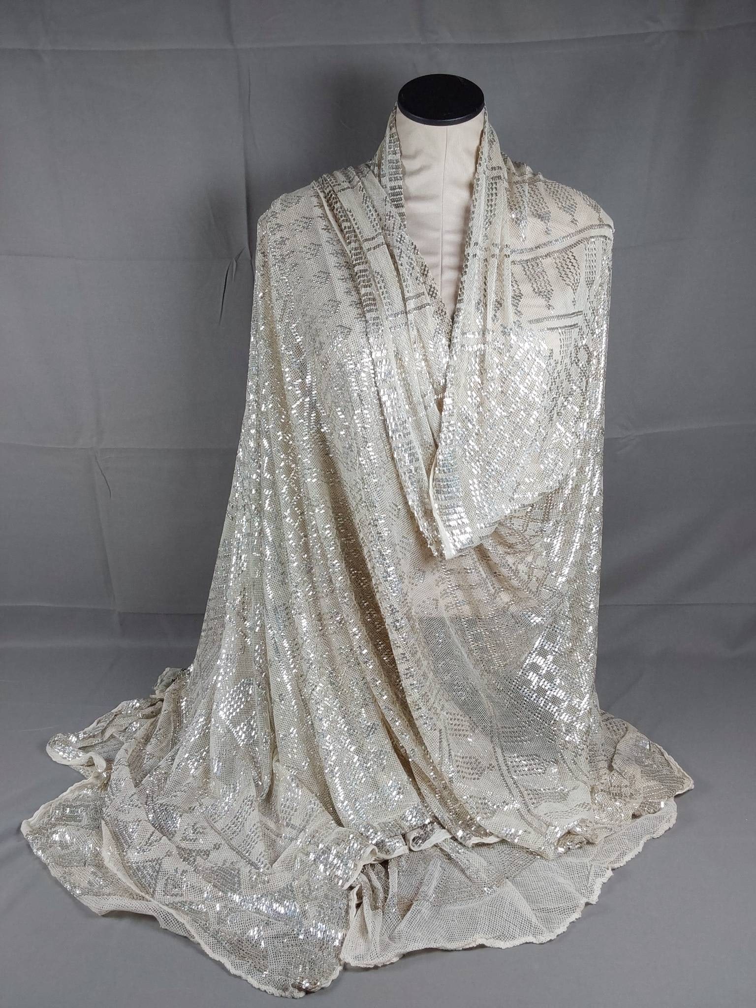 Vintage Silver Assiut Shawl: Vintage Assiut shawl crafted with off-white cotton mesh and metallic embellishments. Features small silver strips woven in a geometric pattern, showcasing skilled craftsmanship. The shawl has a rolled