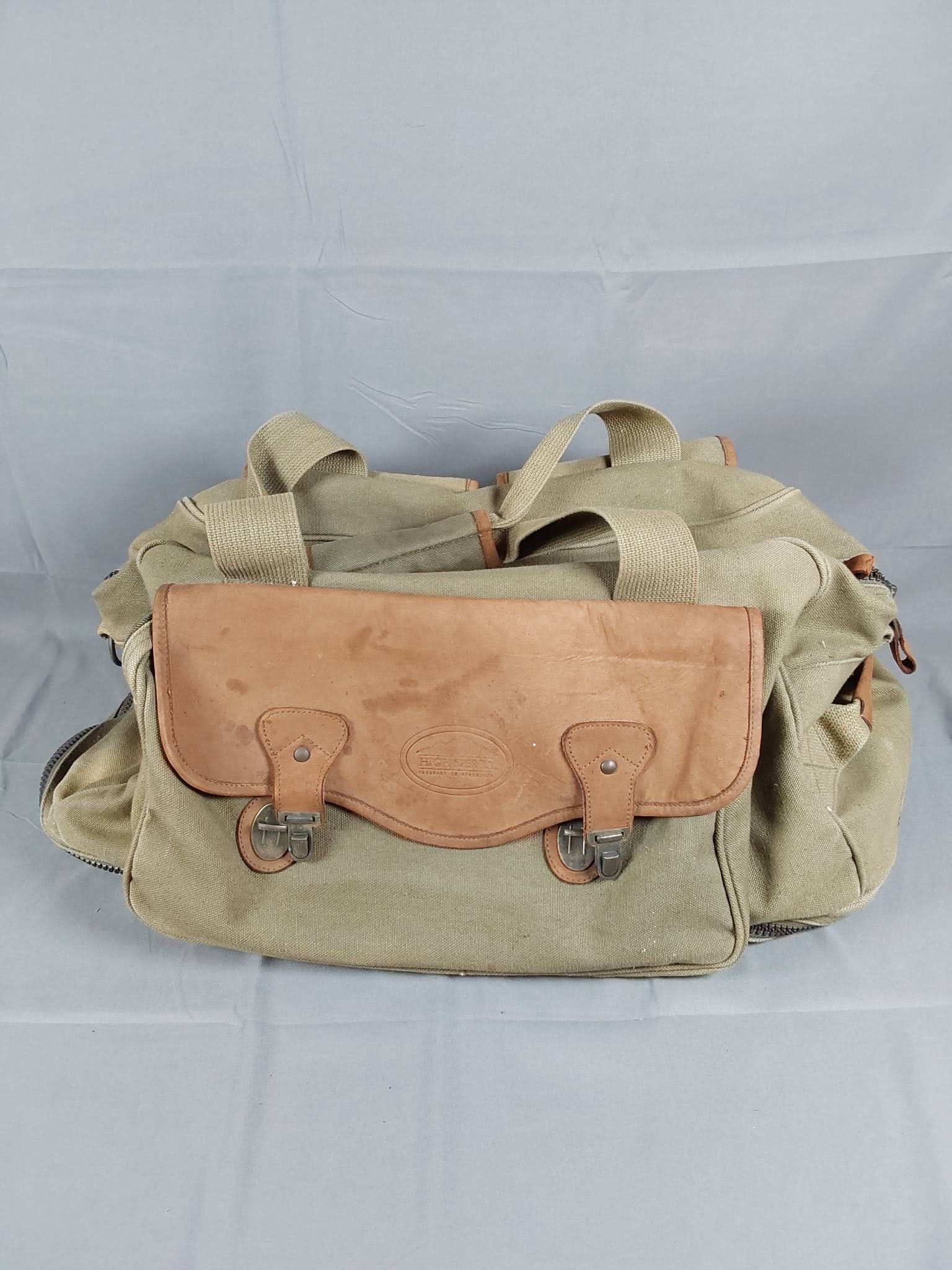 Vintage High Sierra Olive Canvas Duffle Bag: Vintage High Sierra olive green canvas duffle bag featuring brown leather trim and accents. Designed with functionality in mind, it includes a zippered bottom compartment for shoes, two large cargo po