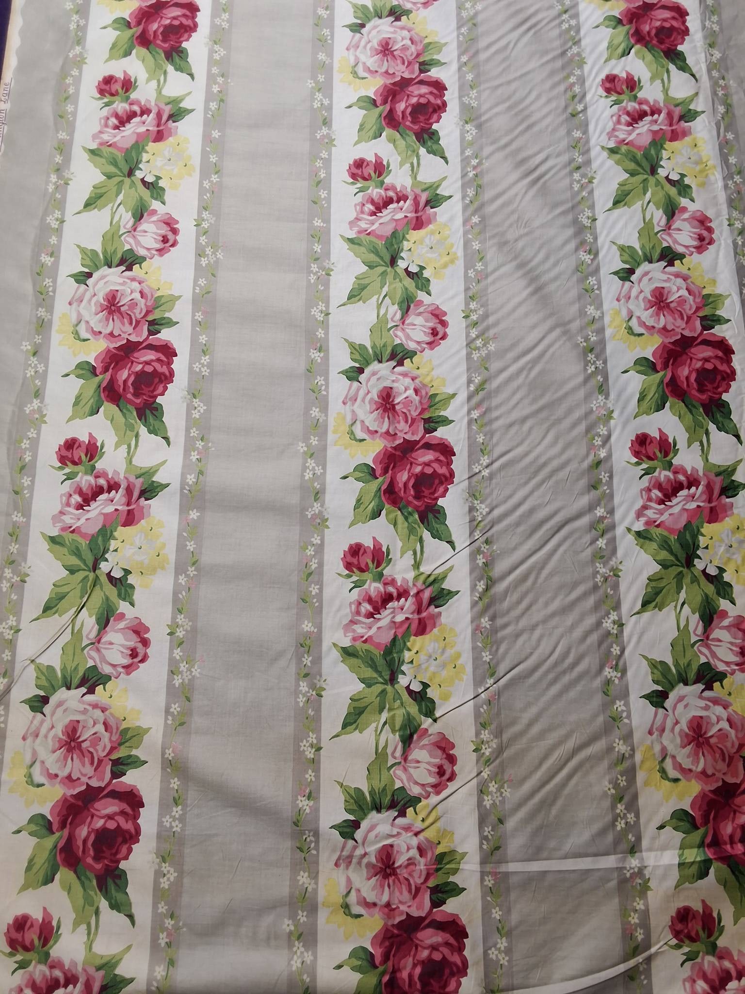 A Waverly Bonded Fabric - Arlington Lane: Vintage cotton home decor fabric measuring 36in by 276in featuring alternating light grey and white stripes. The white stripes showcase red and pink roses with yellow flower clusters alongside hues of