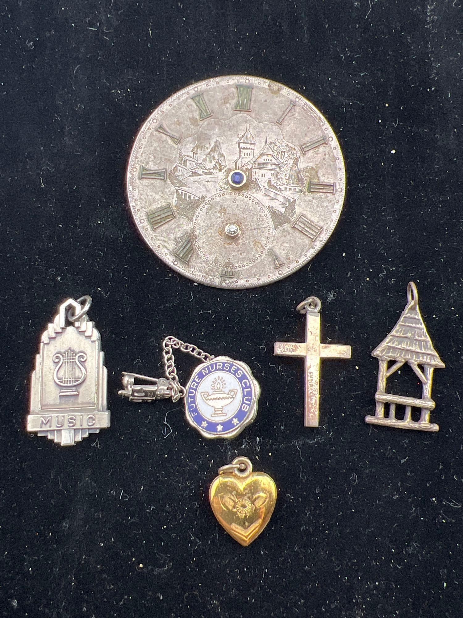 Pins and Pendants: Sterling fraternal pin, sterling cross pendant, silver shack pendant, sterling music pendant, sterling heart locket with gold wash and an unmarked silver engraved clock face brooch 1.5” in diame