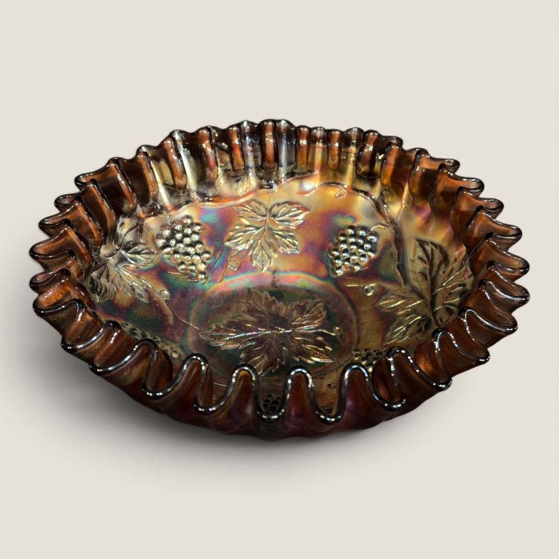 Antique Amethyst Carnival Glass Ruffled Bowl in a Grape and Leaf Pattern: An antique carnival glass bowl featuring an amethyst base glass that appears deep purple to near-black depending on light exposure. The bowl is constructed from pressed glass with a hand-crimped,