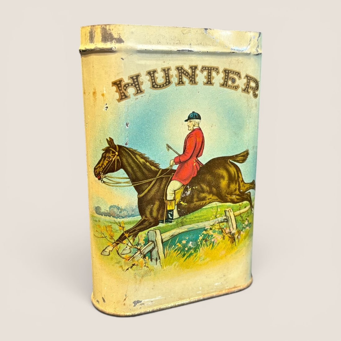 Antique Hunter Brand Lithographed Metal Cigar Tin: A vintage lithographed metal cigar tin produced for the Hunter brand. This pocket-sized tin features a flattened oval cross-section. The front panel is decorated with a multi-color lithograph