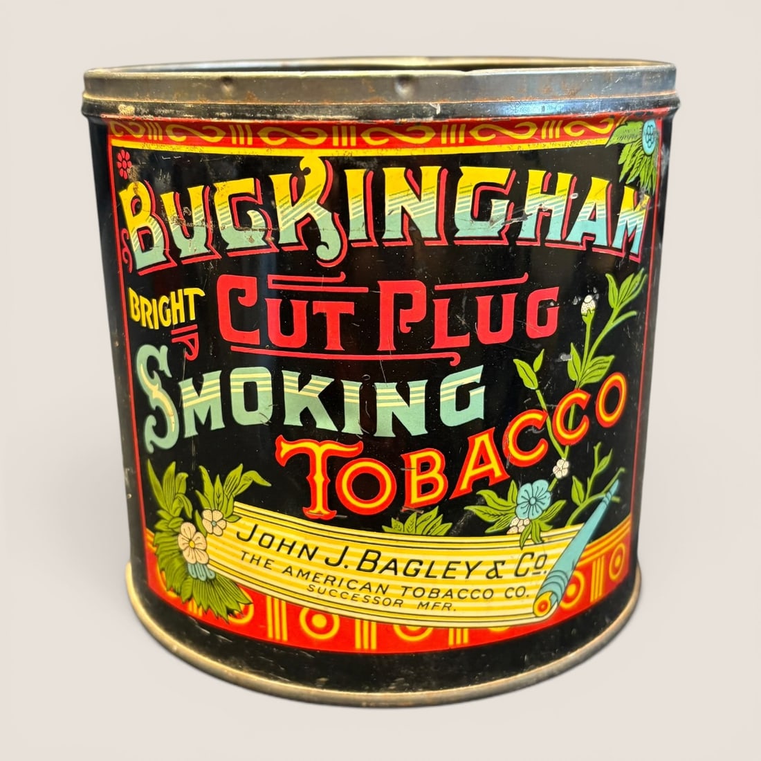 Antique Buckingham Bright Cut Plug Lithographed Tobacco Tin by John J. Bagley and Company: A cylindrical tobacco advertising tin manufactured by John J. Bagley and Company and The American Tobacco Company, Successor Mfr. The tin features a lithographed design on a black ground with vibrant 