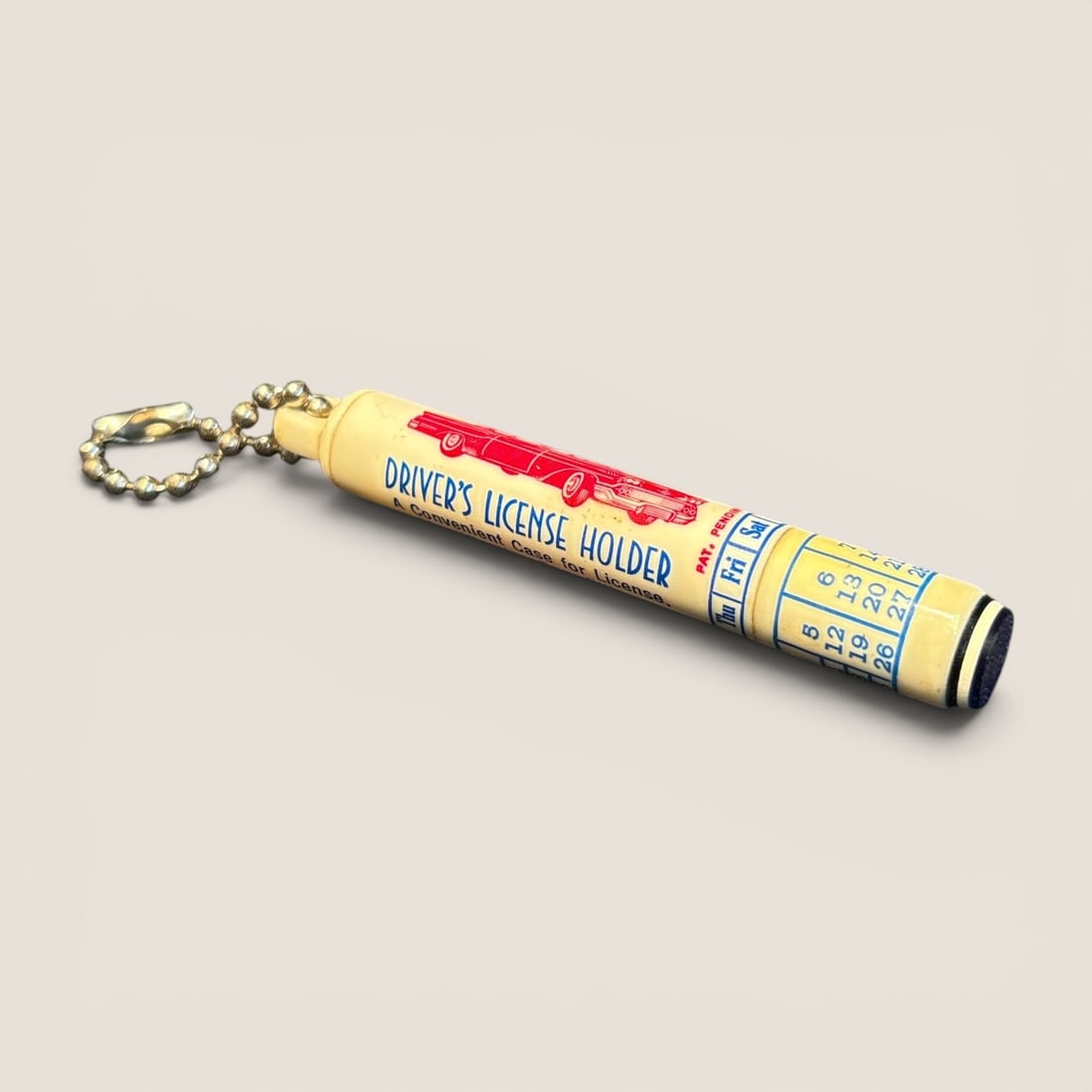 Mid-Century Promotional Plastic Storage Keychain with Perpetual Calendar: A mid-century promotional cylindrical storage keychain, marketed as a multi-purpose holder. The cream-colored plastic body features blue and red printed graphics. One side displays a red sedan above