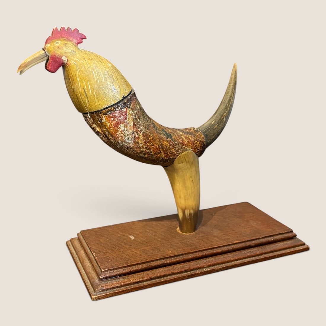 Folk Art Carved Wood and Animal Horn Rooster Sculpture on Tiered Base: This folk art sculpture depicts a rooster in a forward-leaning stance, constructed from a combination of natural animal horn and carved wood. The head and neck are hand-carved from wood and finished w