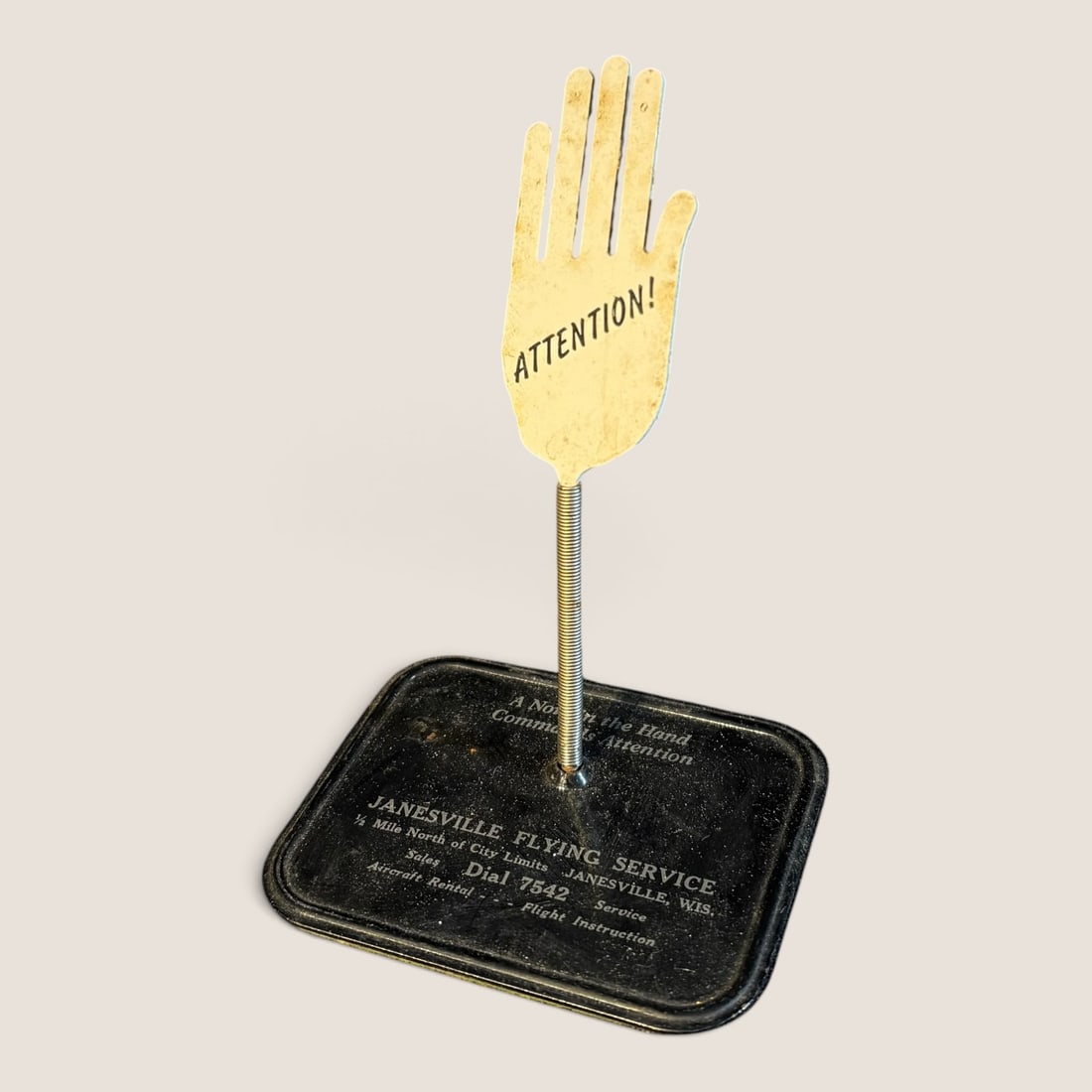 Vintage Janesville Flying Service Advertising Spring Hand Memo Clip: This is a mid-twentieth-century desktop advertising memo holder constructed of metal. The item features a rectangular metal base with rounded corners finished in black enamel. A vertical silver-toned 