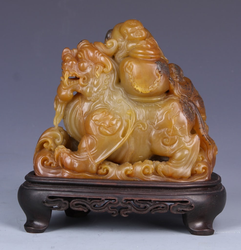 Chinese Shoushan Foo Lion: China, Shoushan style stone carving of foo lion, in reclining position, with detailed face, and two figures on his back, with carved wooden base. Height 5 1/2 in., Length 5 1/2 in.