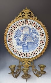 An antique Japanese Imari porcelain plate Mounted with gilt brass as candle holder: An antique Japanese Imari porcelain plate likely from the 19th century, displayed in an ornate gilt brass or bronze frame with a stand that includes three candle holders. 15” H and plate is 10&r
