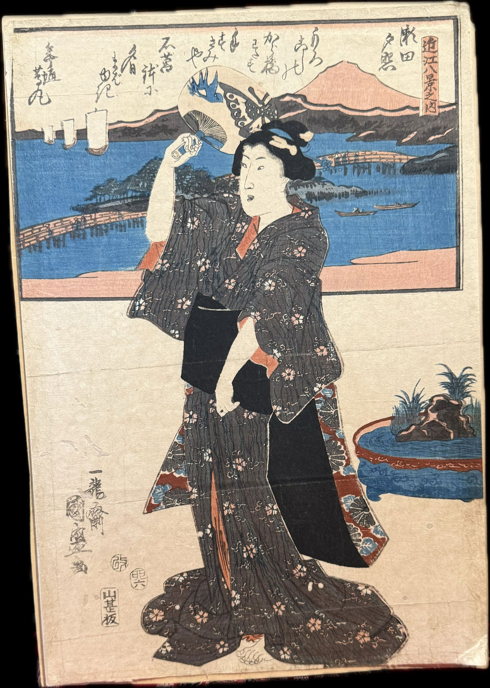 Antique Japanese woodblock print Utagawa Kunisada (1786-1864): Antique Japanese woodblock print is a ukiyo-e design by the artist Utagawa Kunisada, active during the Edo period. The artwork features a bijin (beautiful woman) holding a fan, with a landscape scene