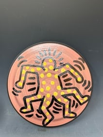 A porcelain plate featuring a dancing figure with a signature of P. Harings 87: A porcelain plate featuring a dancing figures in yellow and black on a pinkish with a signature of P. Harings, 87, 8” D