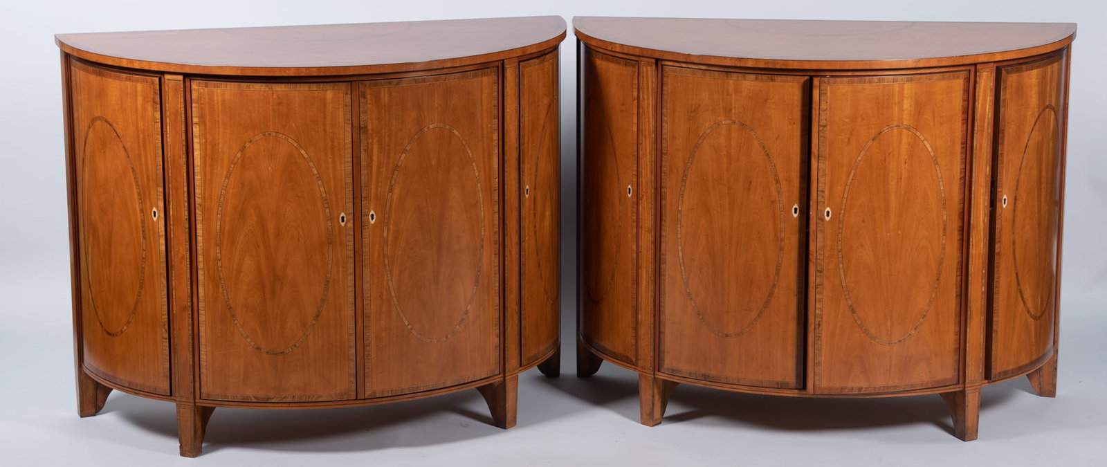 A Pair Of Baker Demi Lune Consoles: A Pair Of Baker Demi Lune Consoles The top having inlay and crossbanding above four hinged doors with oval inlaid pattern and ribbon inlaid borders supported on four tapering supports. 31 1/2 x 42 x 2