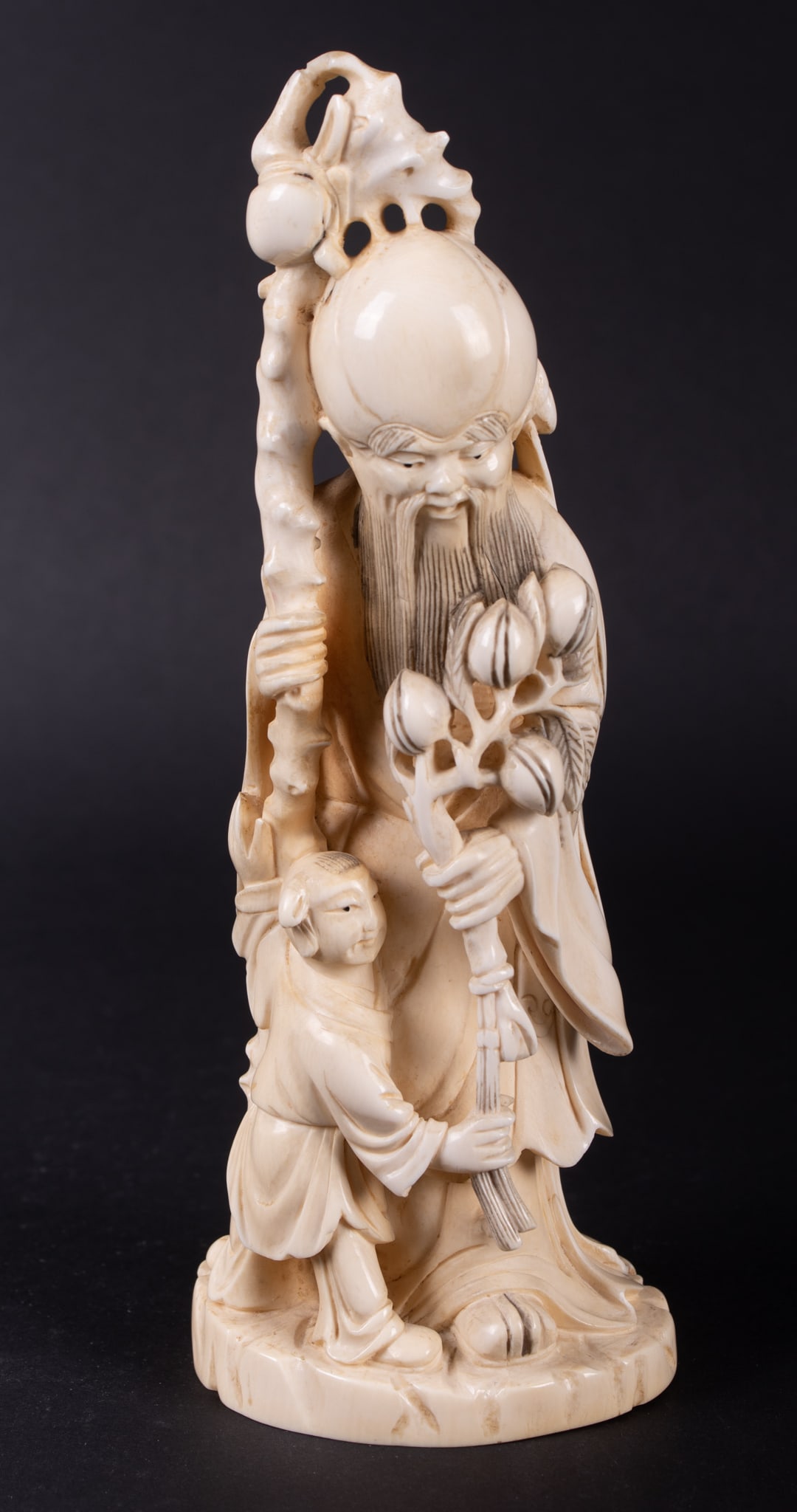 A Chinese Bone Carving of Shou Lao: A Chinese Bone Carving of Shou Lao China, a carving depicting Shou Lao, symbolic of longevity, with branches of peach clusters and attended by a student. 8 3/4 inches tall