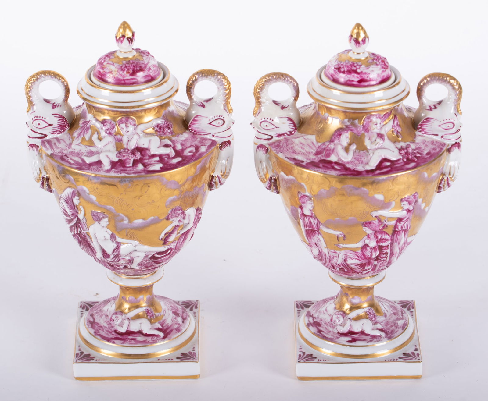 A Pair Of Antique Capodimonte Lidded Urns: A Pair Of Antique Capodimonte Lidded UrnsAn unusual pair of well decorated Capodimonte urns, each having classical scenes and dolphin handles with red and gold gilt highlights.Provenance: The Estate o