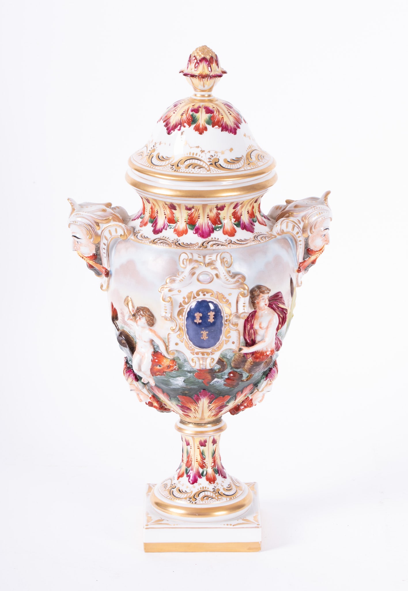 A Capodimonte Lidded Porcelain Urn: A Capodimonte Lidded Porcelain UrnAn elaborate bolted urn, the body with highly raised figures, sea nymphs and cupids; the double handles masks of Neptune. Bears blue N-Crown mark to base.