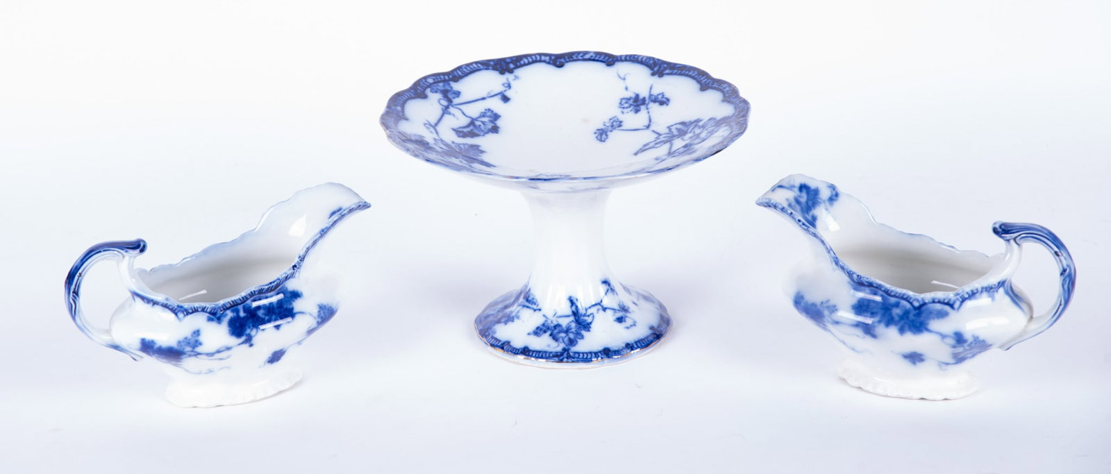 A Pair of Antique Staffordshire Flow Blue Gravy Boats and a Pastry Compote: England, circa 1890, a pair of Virginia pattern gravy boats by John Maddock and Sons (one with crazing and a hairline near the handle). Paired with a similar grapevine pattern footed compote in the Lo