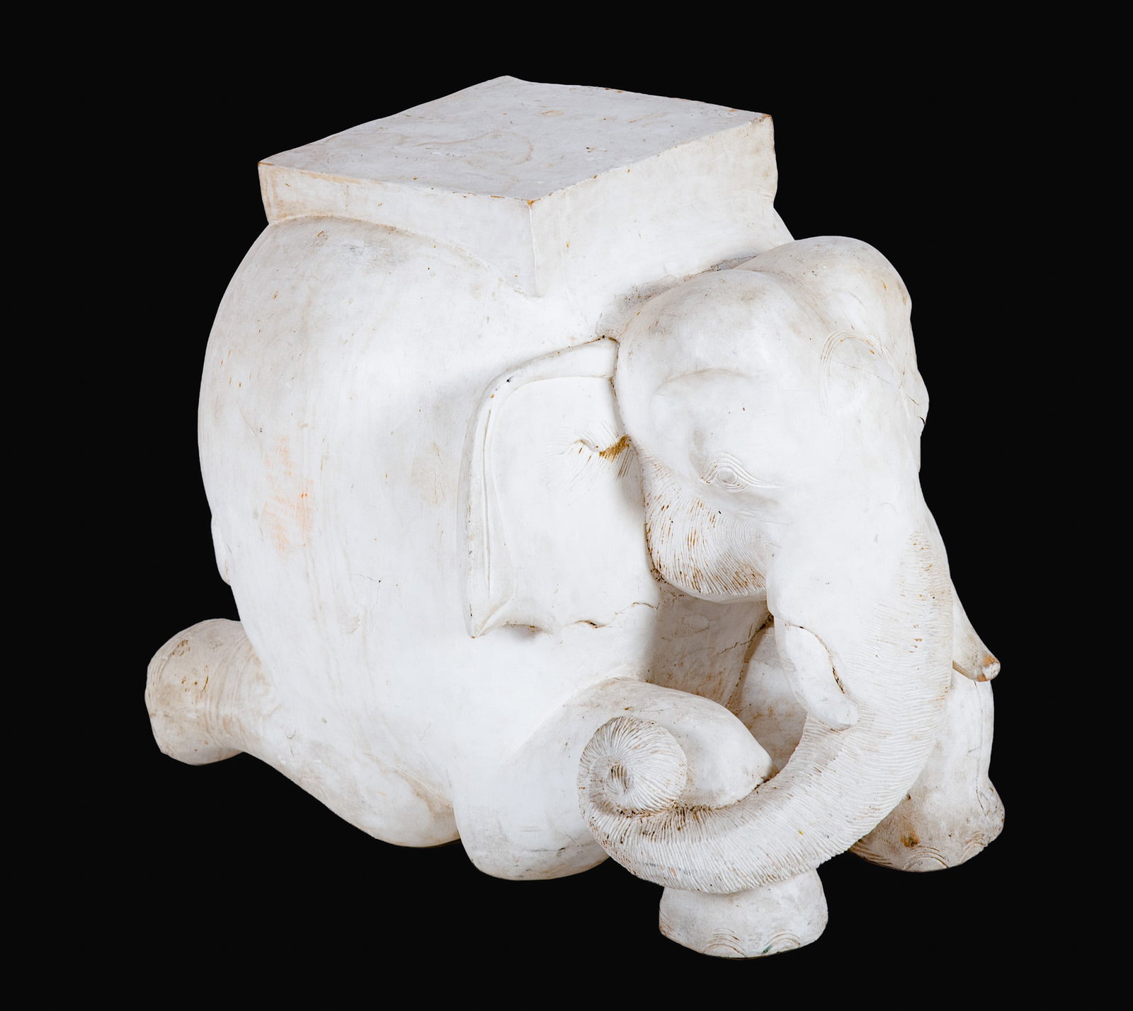 A Large Hand Carved Crouching Elephant: A Large Hand Carved Crouching Elephant The white painted elephant figure has a square pedestal top, and could be used as a table base. Height: 23 inches; Width: 19 inches; Depth: 26 inches
