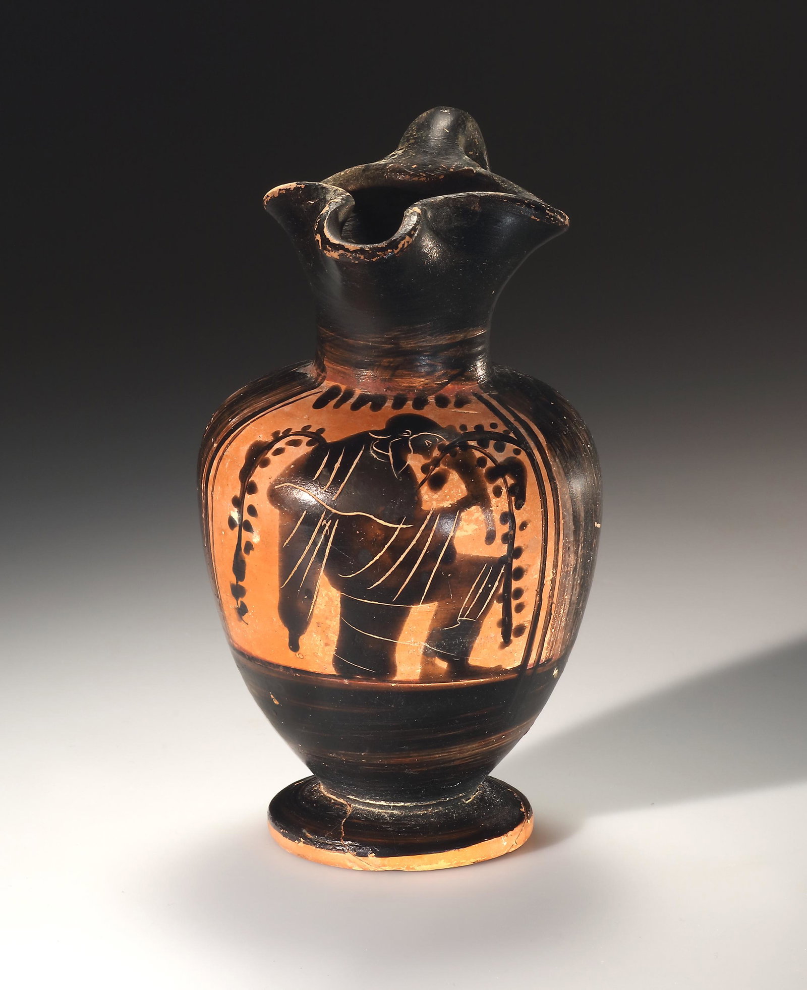 A Black Figure Oinochoe with Dionysos: Attic 525-475 B.C..Pale orange clay with fine mica and red slip, black glaze, added white. H. max. 13 cm.Black-glazed wine jug with trefoil mouth. In the reserved picture field, Dionysus stands betwee