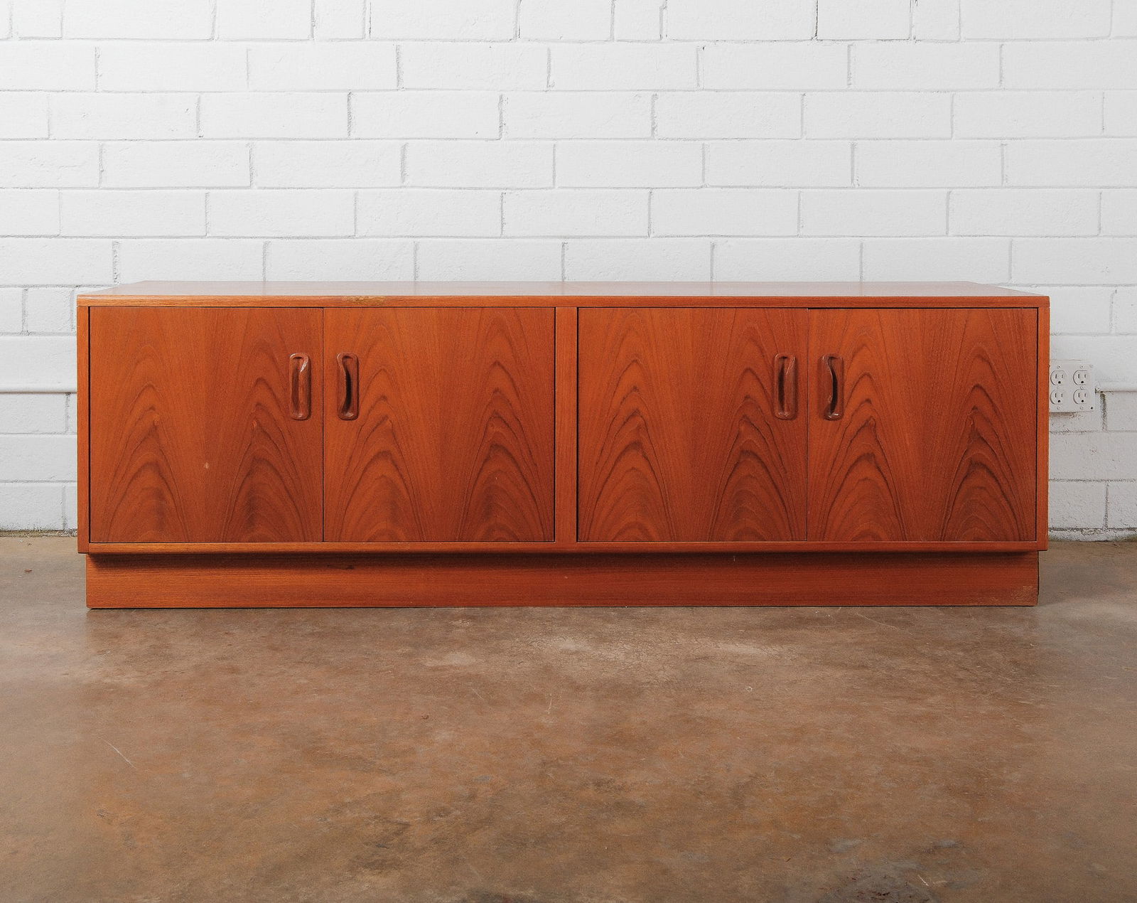 Mid-Century Modern Low Credenza/Sideboard: A sleek low profile mid-century modern credenza featuring four flat front doors in a warm wood veneer with recessed vertical pulls. The piece rests on a plinth base, emphasizing its clean lines and mi