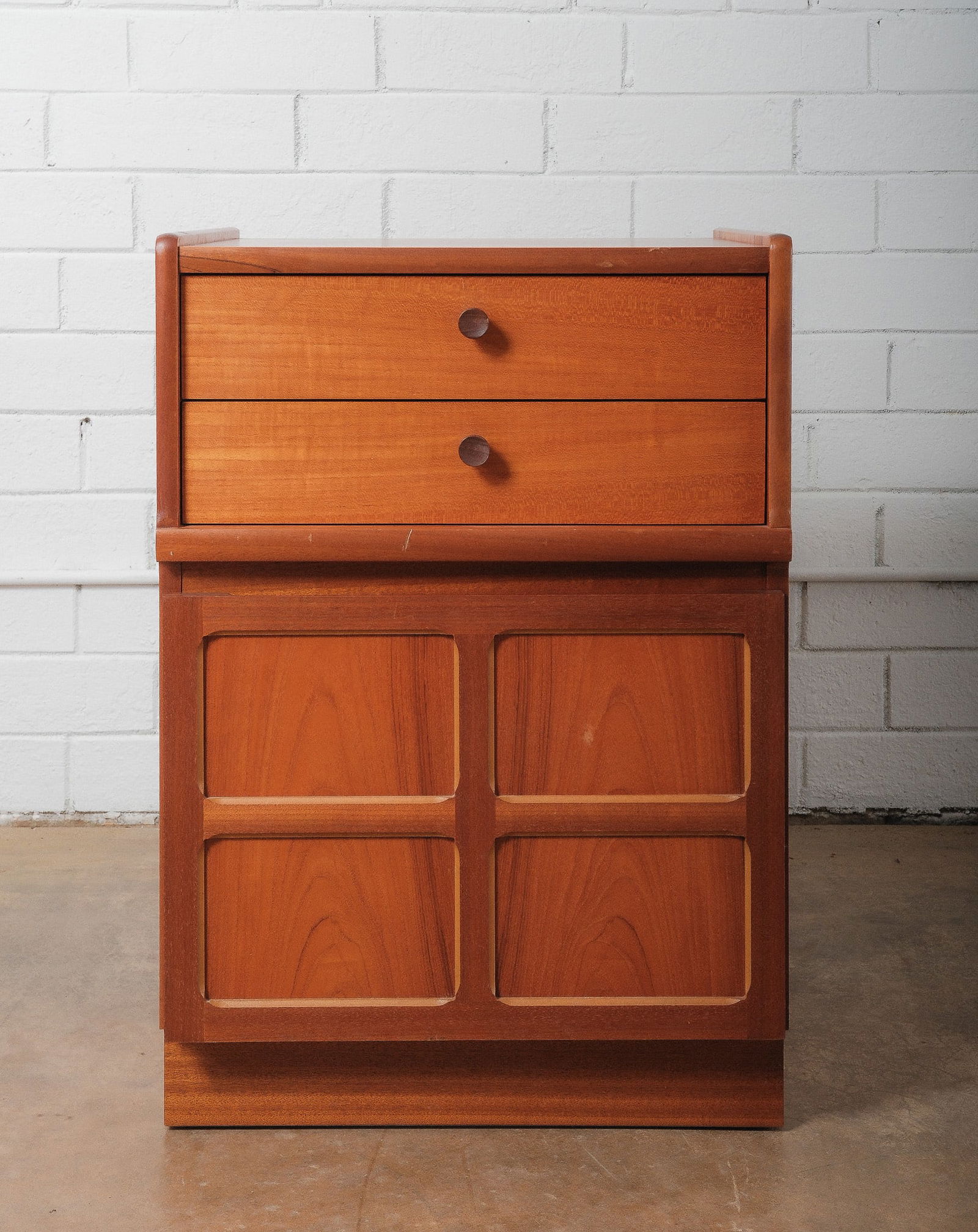 Mid-Century Modern Two Drawer Cabinet with Lower Doors: A petite mid-century modern cabient featuring two upper drawers over a double-door lower cabinet. Constructed in warm toned wood veneer with recessed panel details and simple round door pulls. Compact