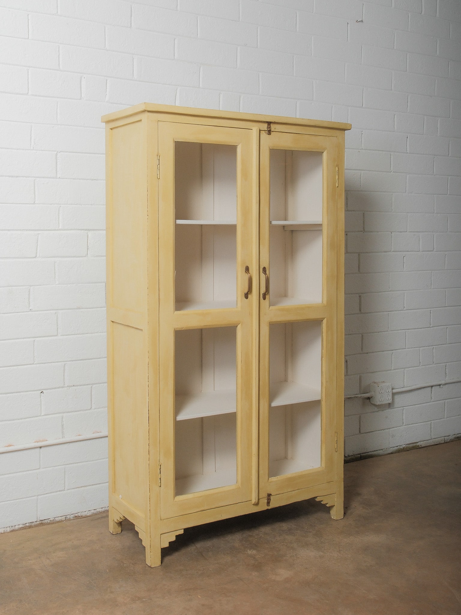 Yellow Painted Glass-Door Cabinet: Charming painted cabinet featuring double glass doors with original warm brass/bronze pulls and a soft butter-yellow finish. The piece showcases a classic country construction with framed glass panels