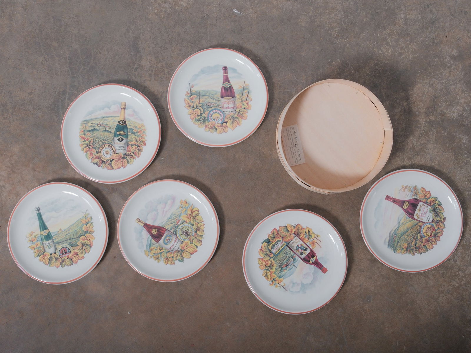 Set of 6 Vintage Cheese Plates With Original Box: Set of six decorative porcelain plates, each featuring a different wine bottle vignette framed by grape vines and scenic vineyard landscapes. Includes the original round wooden presentation box. Charm