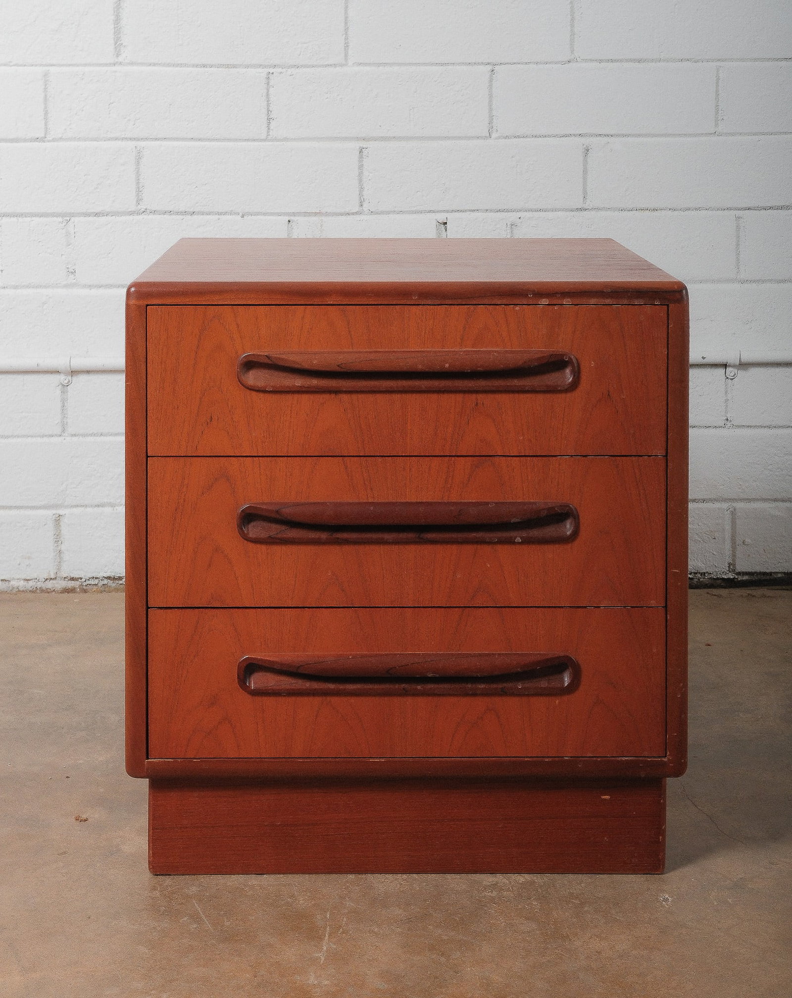 MCM Small Chest of Drawers: This mid-century modern nightstand features a compact three-drawer design with sculpted integrated pulls that emphasize its clean, minimalist aesthetic. Finished in a warm wood tone, the piece is rais