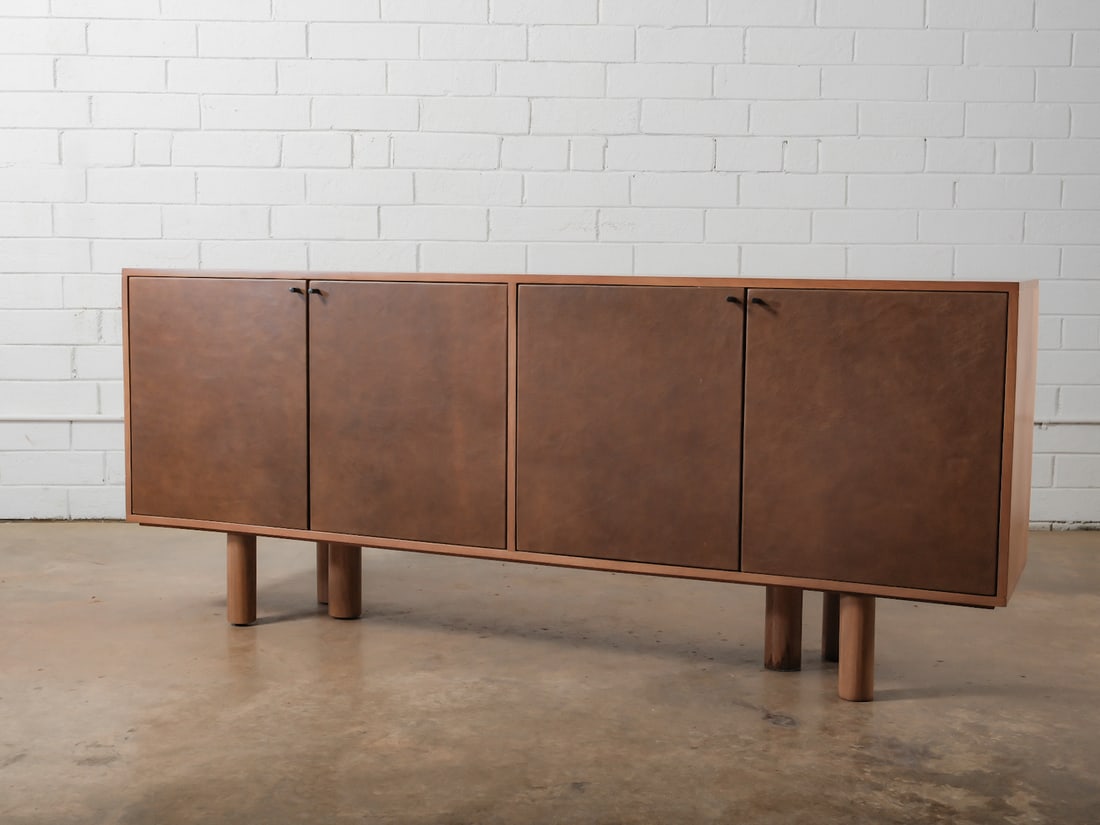 Modern Storage Credenza with Leather Doors: A long credenza featuring a clean, minimalist silhouette. The piece rests on cylindrical legs and offers concealed storage behind four flat-front leather finished doors. The warm brown finish highligh