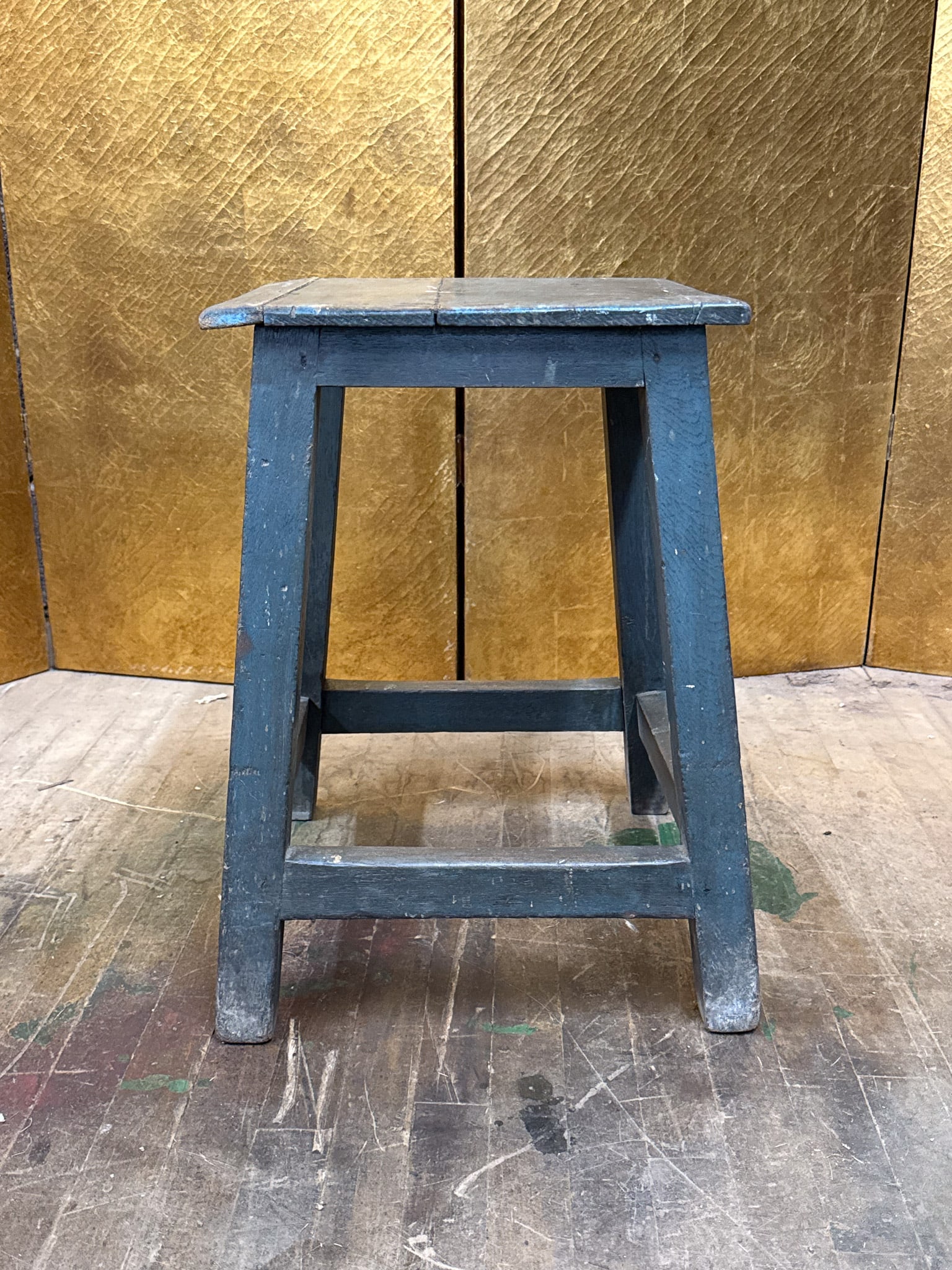 Vintage Painted Wooden Stool: Vintage wooden stool featuring a sturdy square seat and tapered legs joined by stretchers for added support. Finished in a blue painted surface with visible wear that adds to its rustic character. A