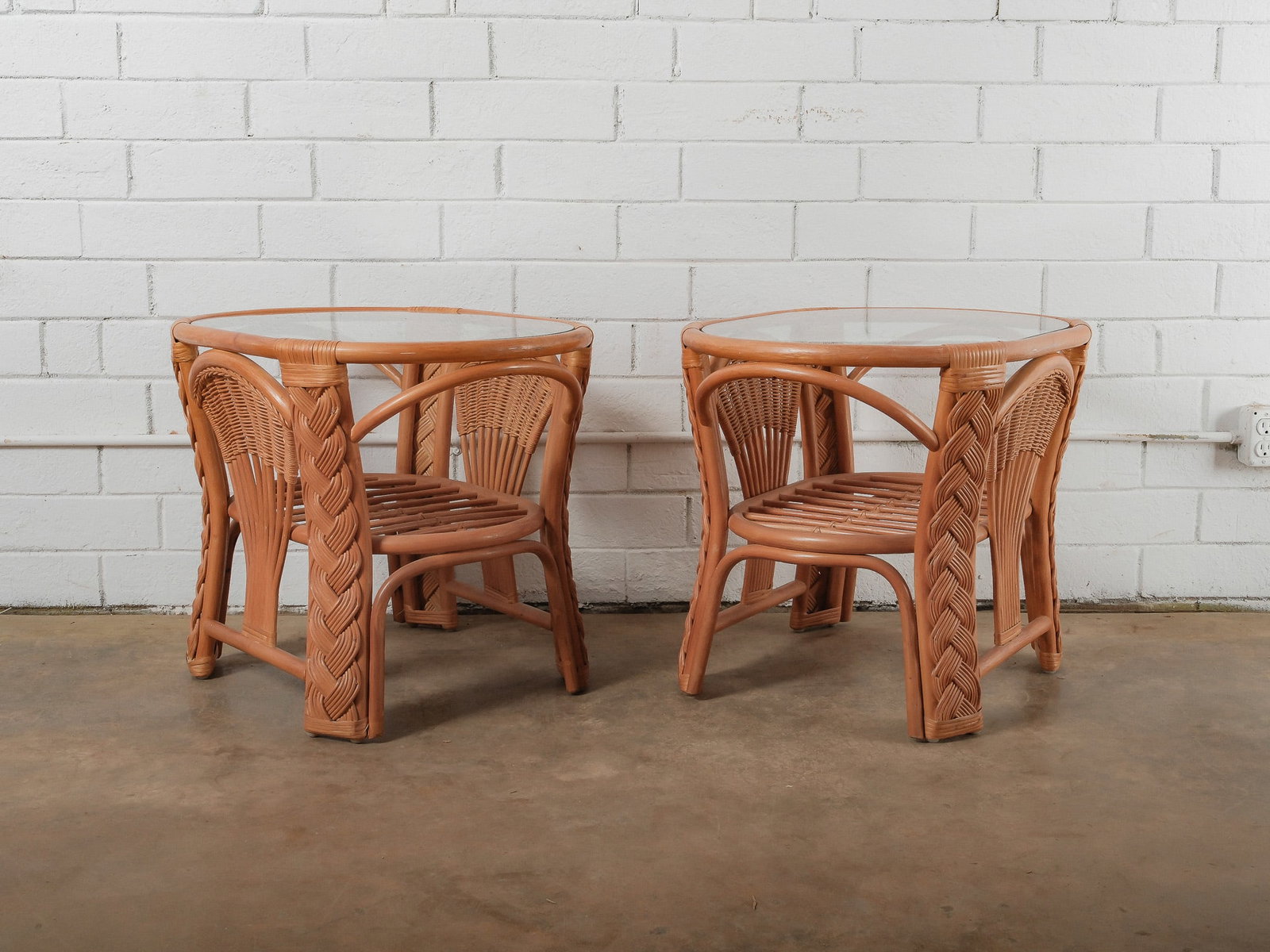 Pair of Rattan Side Tables: Matching pair of rattan side tables, each topped with a circular glass surface. The bases feature woven rattan detailing with braided accents along the legs, offering a sculptural and textural