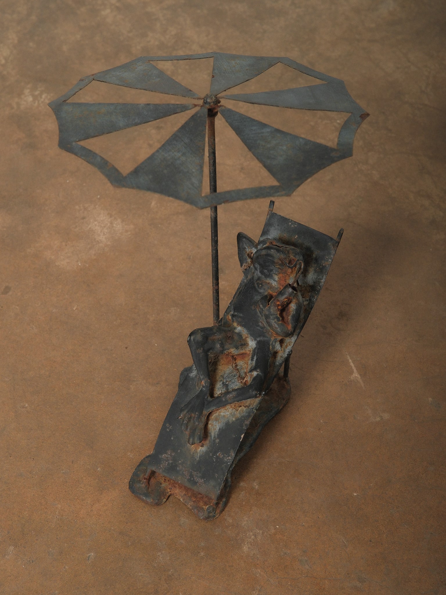 Bronze Frog Sculpture Reclining in Sun Lounger: Decorative bronze frog sculpture depicting a whimsical figure reclining on a curved lounge chair beneath a stylized umbrella. The piece features a rustic patinated finish with sculptural metal constru