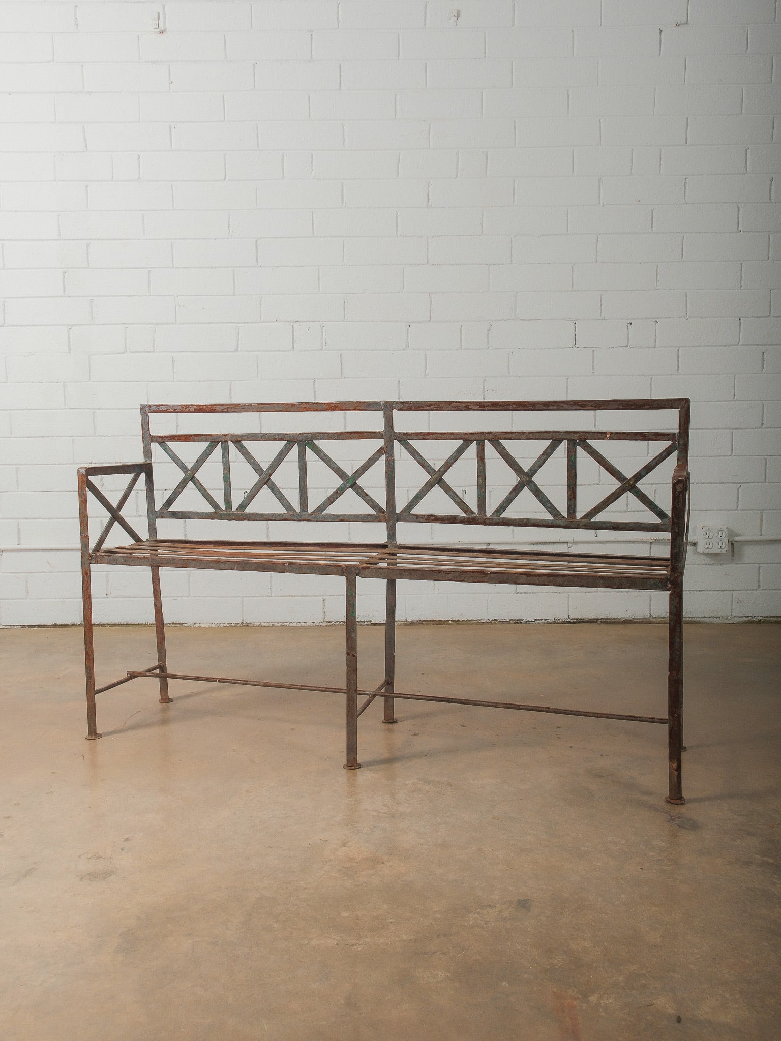 Industrial Iron Garden Bench with X-Form Back: Metal garden bench featuring a decorative X-form backrest and slatted seat. The iron frame shows natural rust and weathered patina consistent with outdoor use. Ideal for garden seating, patio use, or