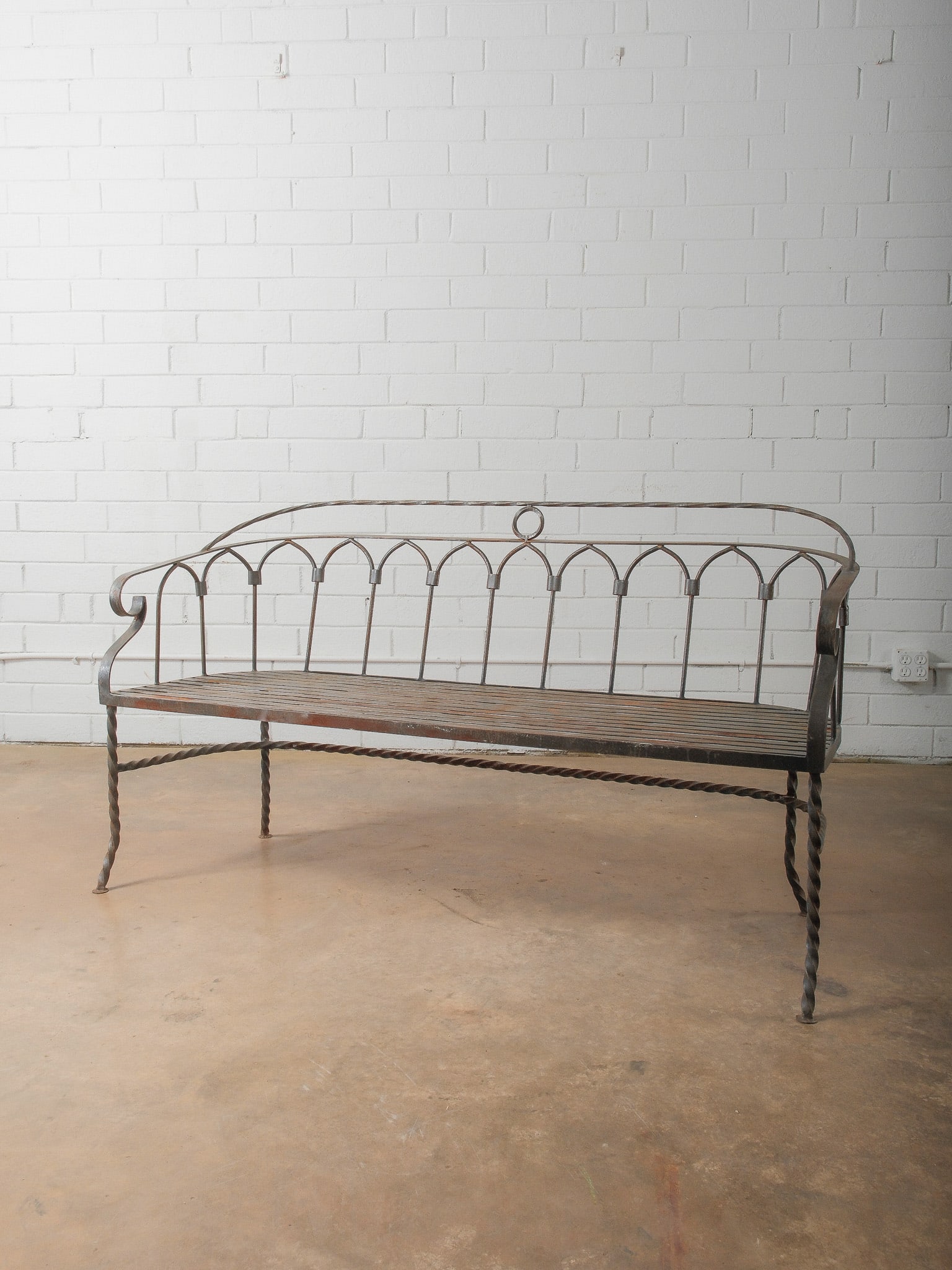 Wrought Iron Garden Bench with Arched Back: Decorative wrought iron garden bench featuring a graceful arched back with repeating Gothic-style motifs and central ring detail. The seat is constructed of slatted iron, supported by twisted iron leg