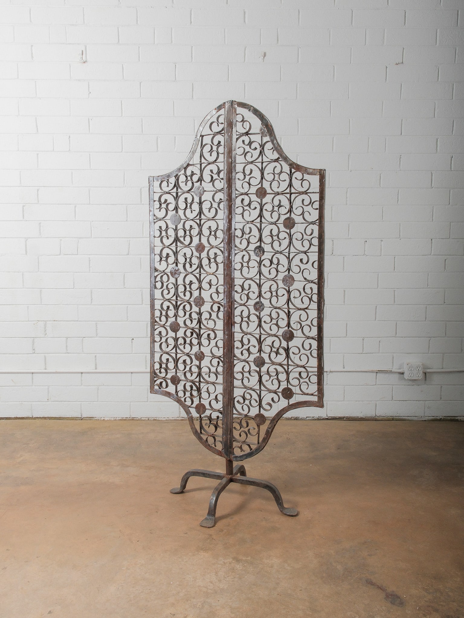 Decorative Wrought Iron Standing Screen on Pedestal Base: free standing double-sided wrought iron screen featuring intricate scrollwork panels with round medallion accents and an arched top silhouette. Mounted to a central vertical support with four-footed