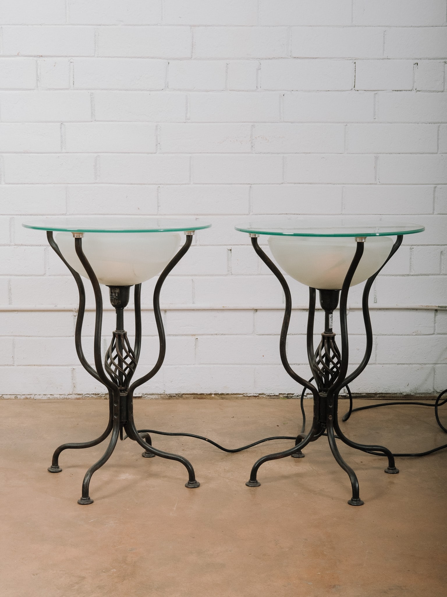 Pair of Illuminated Glass and Metal Accent Tables: Pair of illuminated accent tables featuring round glass tops supported by sculptural black metal bases with integrated frosted glass uplights. The decorative metalwork and built-in lighting create a