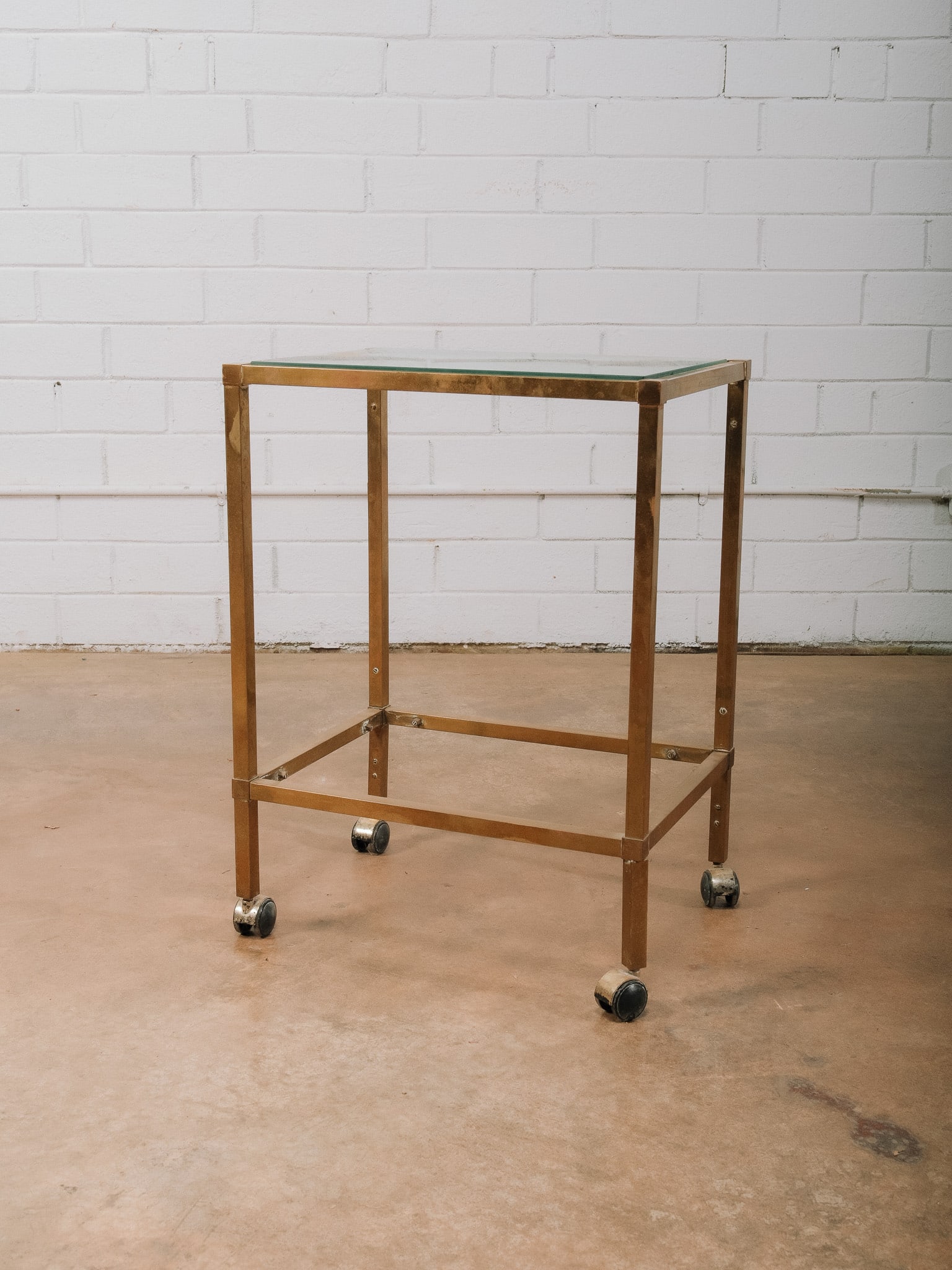 Rolling Brass and Glass Bar Cart: Vintage rolling bar cart featuring a brass-finished metal frame with a glass top and lower shelf mounted on four casters for easy mobility. Its clean, minimalist design works equally well as a bar