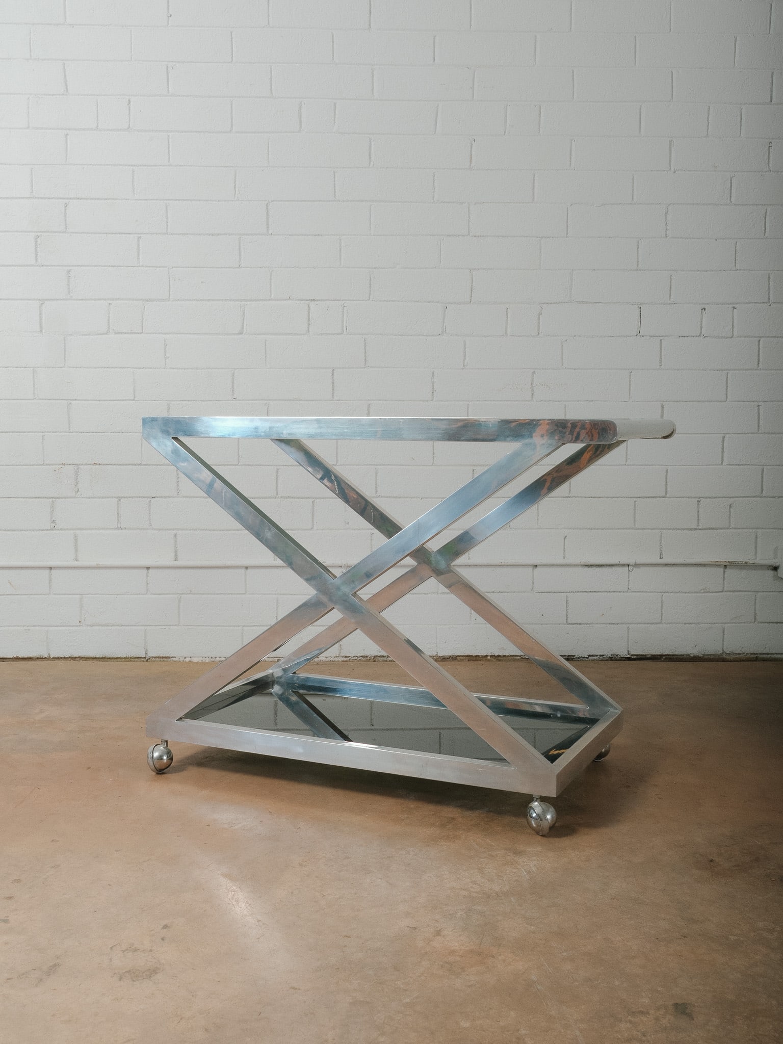 Modern Chrome and Glass Bar Cart on Casters: Modern bar cart featuring a polished chrome frame with a geometric X-form design and two smoked glass shelves. The upper tier includes a built-in handle for easy maneuverability, while the lower