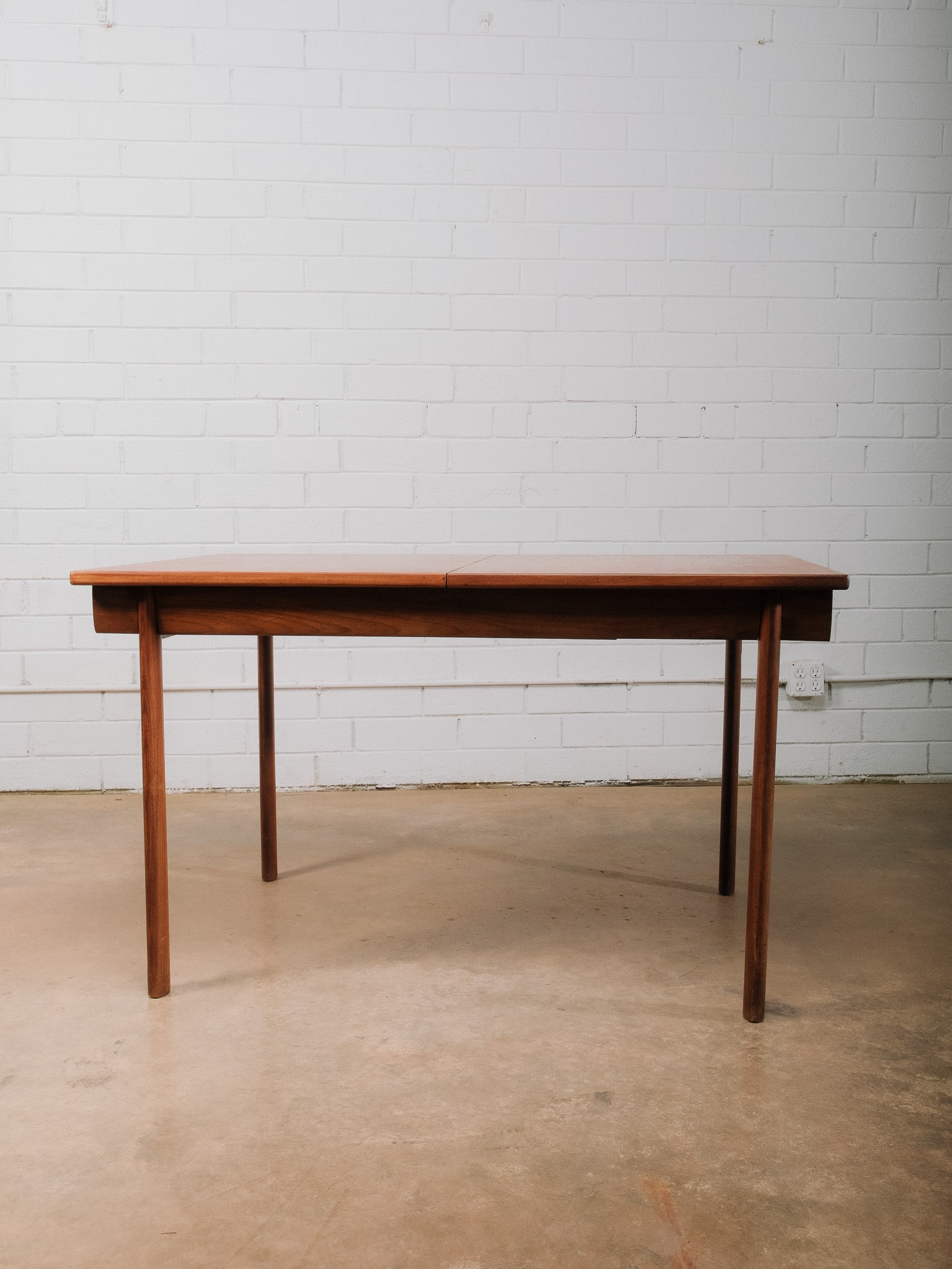 MCM Dining Table: Mid-century modern dining table featuring clean lines, tapered round legs, and a warm wood finish. The table includes a cleverly concealed self-storing extension leaf, allowing it to expand easily