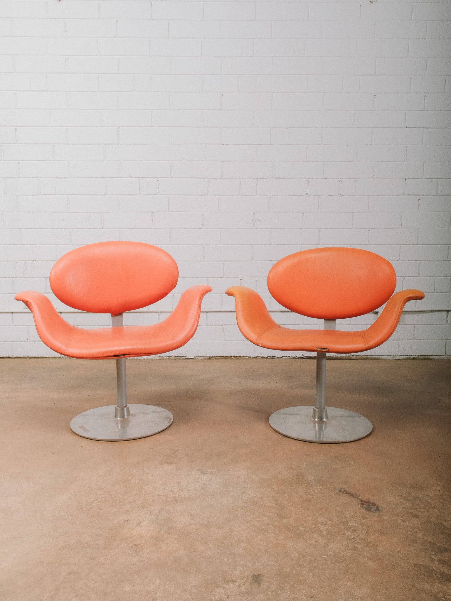 Pair of Mid-Century Modern Orange Swivel Chairs: Pair of mid-century modern swivel lounge chairs upholstered in vibrant orange vinyl with sculptural, space-age styling. Each chair features a rounded backrest, curved integrated arms, and a brushed