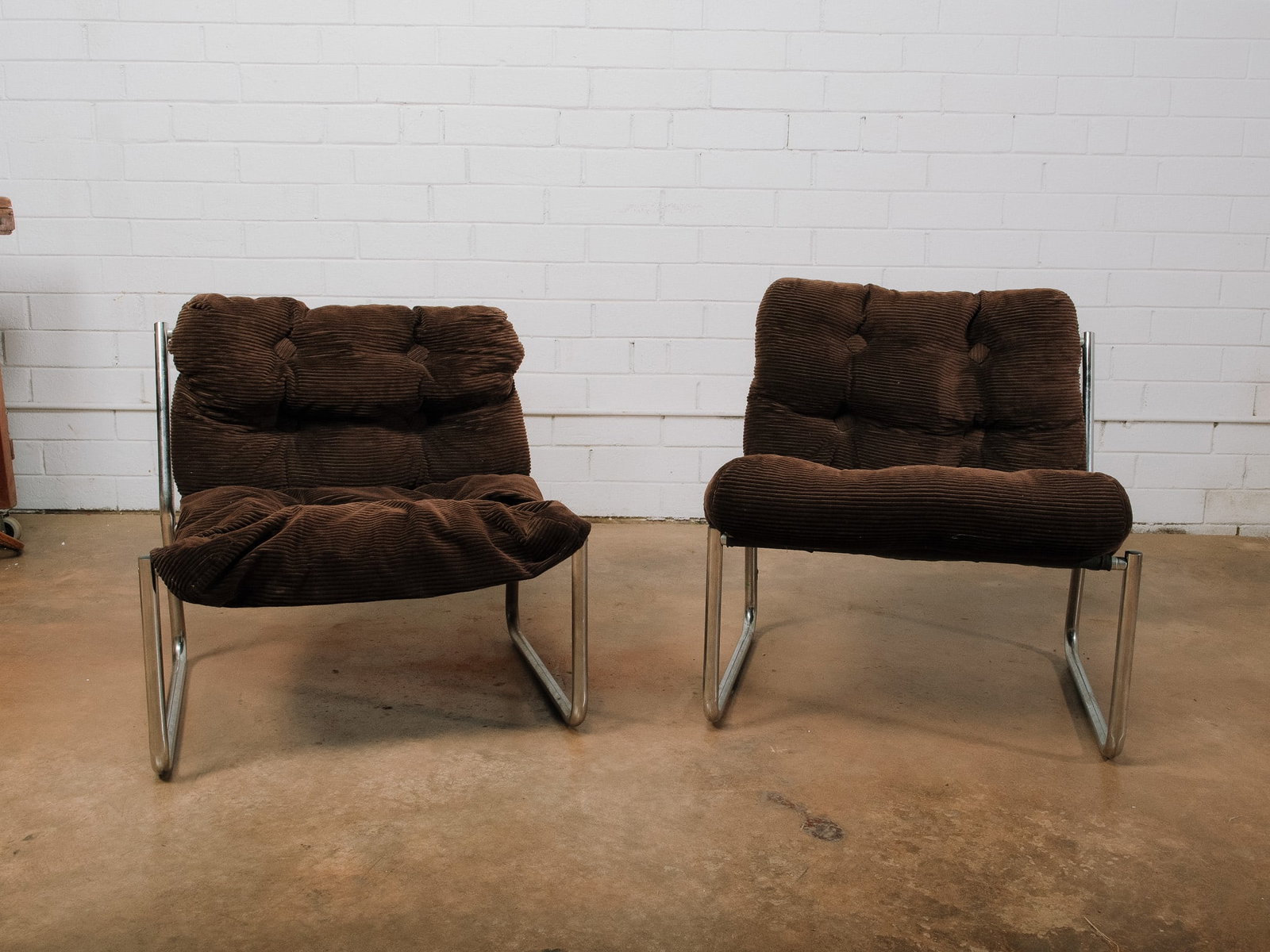 Pair of Chrome Lounge Chairs: Pair of mid-century style lounge chairs featuring tubular chrome frames and oversized tufted corduroy cushions in a rich brown tone. Designed for relaxed comfort, these low-profile chairs make a